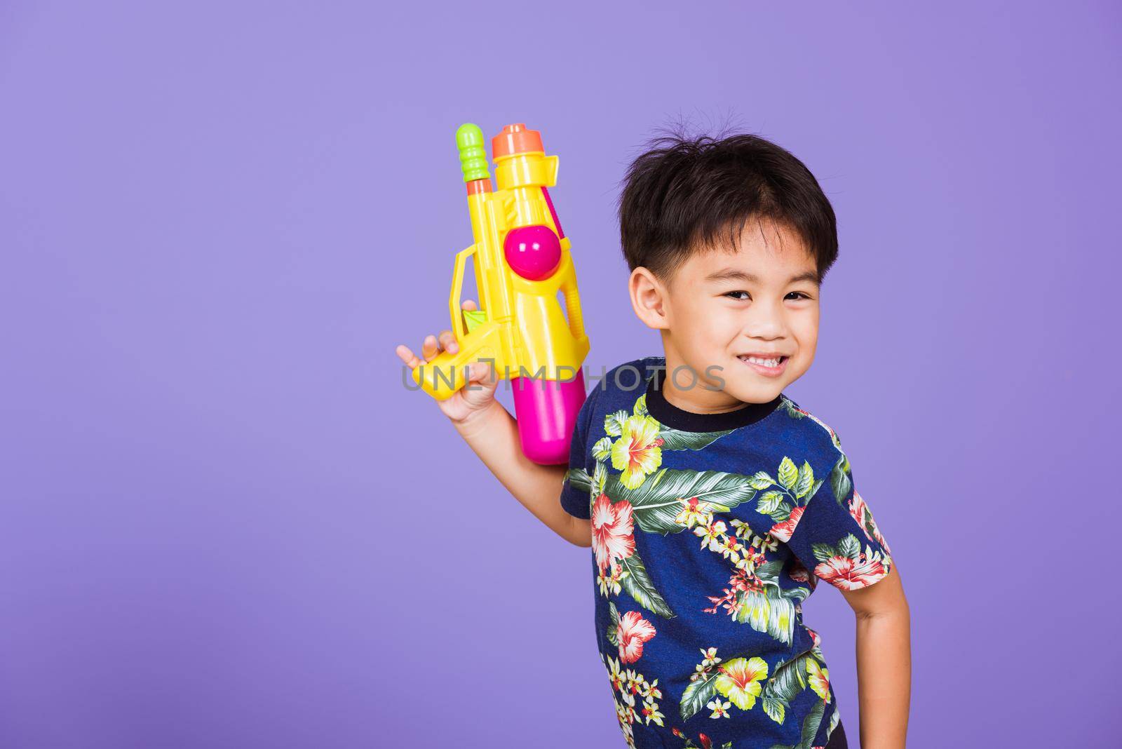 Thai kid funny hold toy water pistol and smiling, Happy Asian little boy holding plastic water gun, studio shot isolated on purple background, Thailand Songkran festival day national culture concept