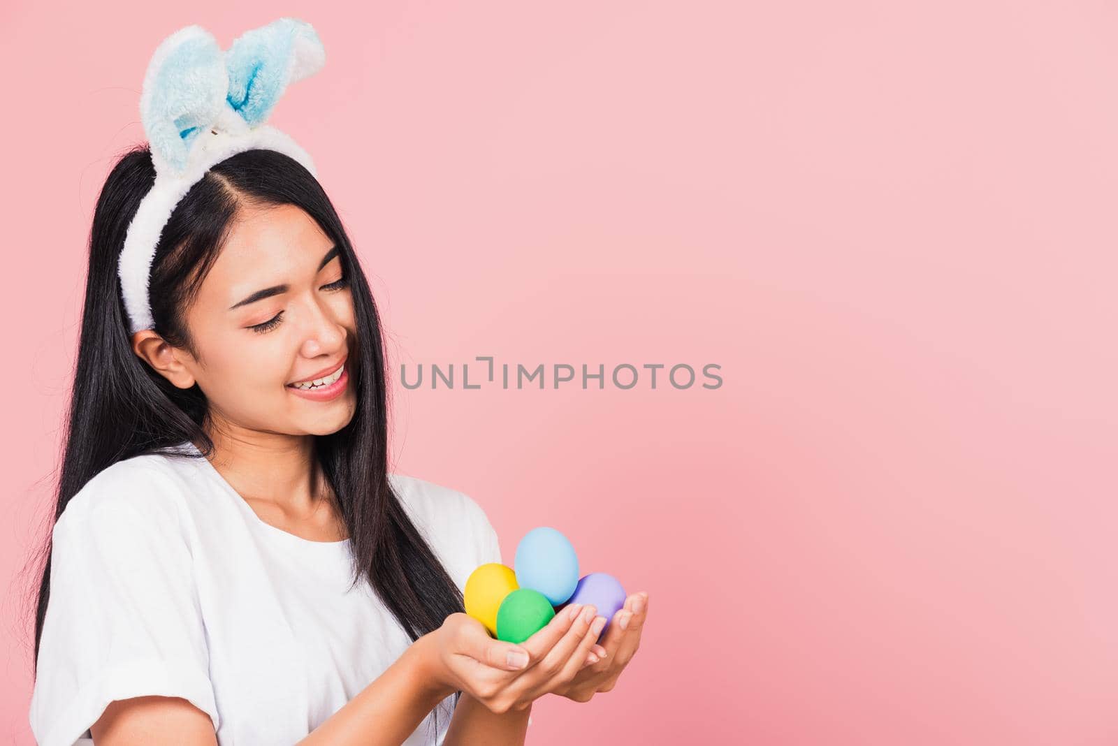 Happy Easter concept. Beautiful young woman smiling wearing rabbit ears holding colorful Easter eggs gift on hands, Portrait female looking at eggs, studio shot isolated on pink background