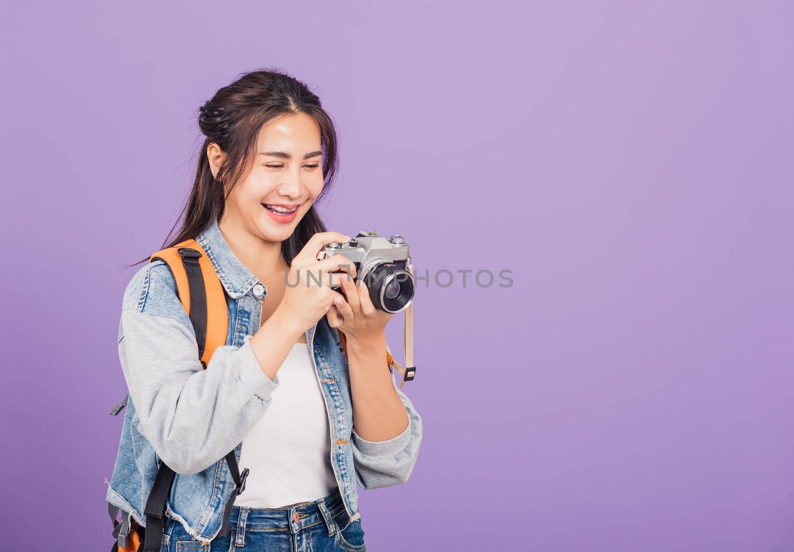 Attractive portrait happy Asian beautiful young woman smiling excited wear denims and bag holding vintage photo camera, female traveler female photographer, studio shot isolated on purple background