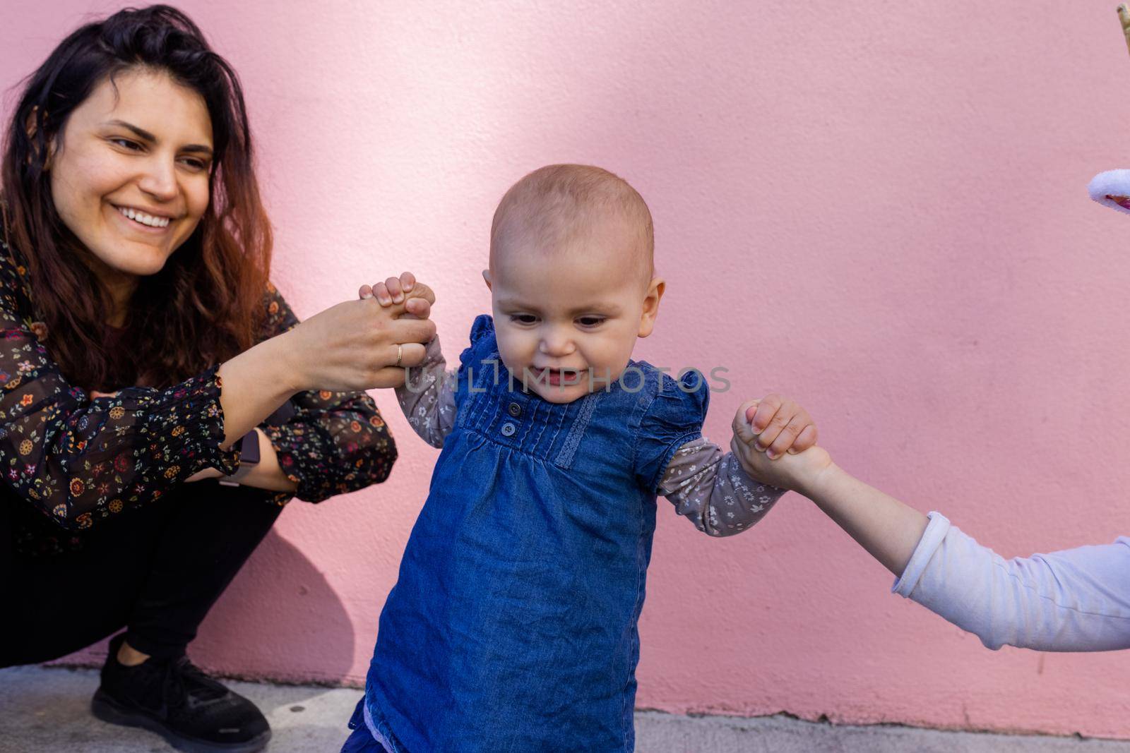 Portrait of woman and little girl holding cute smiling baby by the hands with pink wall as background. Adorable baby standing on sidewalk between her mother and older sister. Happy family outdoors