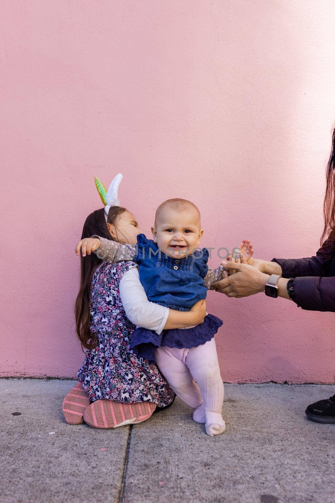 Adorable view of little girl wearing unicorn headband hugging her happy baby sister with pink background. Mother holding cute toddler hand while sitting in front bright pink wall. Lovely kids outdoors