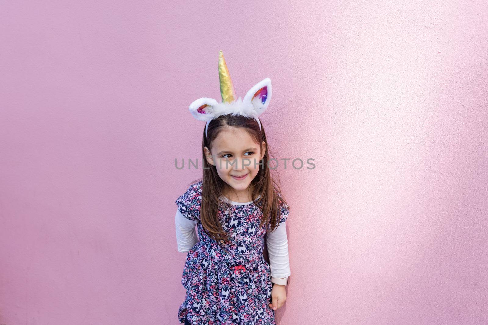 Adorable view of happy little girl wearing unicorn headband with pink background. Portrait of cute smiling child with unicorn horn and ears in front bright pink wall. Lovely kids in costumes