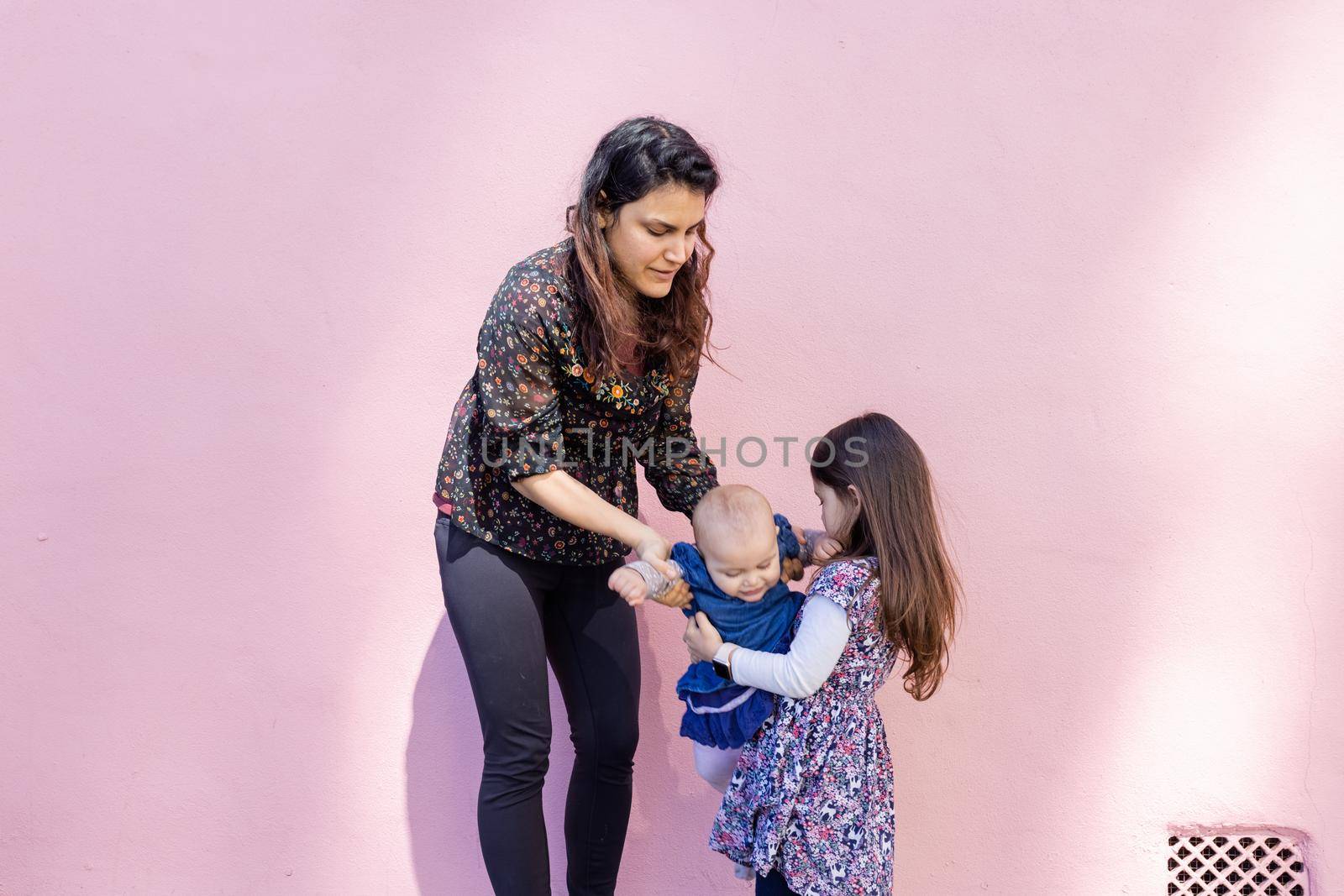Portrait of woman helping little girl to hold cute smiling baby with pink wall as background. Adorable view of baby in arms of her older sister and mother. Happy family outdoors