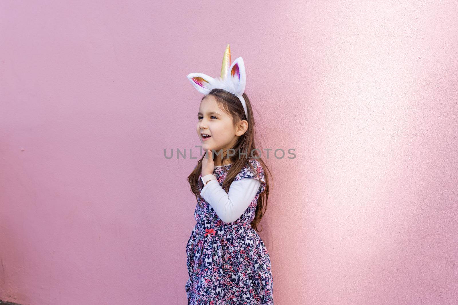 Adorable view of happy little girl wearing unicorn headband with pink background. Portrait of cute smiling child with unicorn horn and ears in front bright pink wall. Lovely kids in costumes