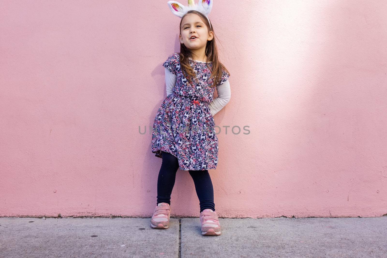 Adorable view of happy little girl wearing unicorn headband with pink wall as background. Portrait of cute smiling child with unicorn horn and ears standing on the sidewalk. Lovely kids in costumes