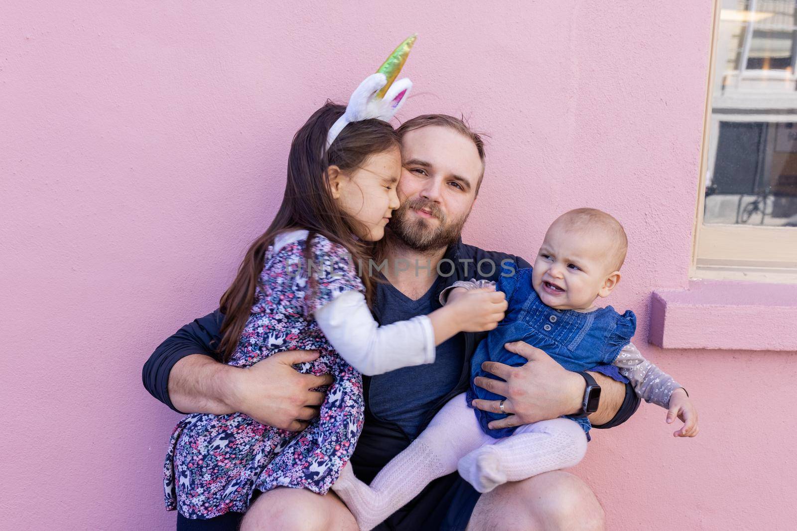 Portrait of young father hugging his adorable baby and older daughter in front of a pink wall. Bearded man in squatting position holding baby and little girl. Happy family outdoors