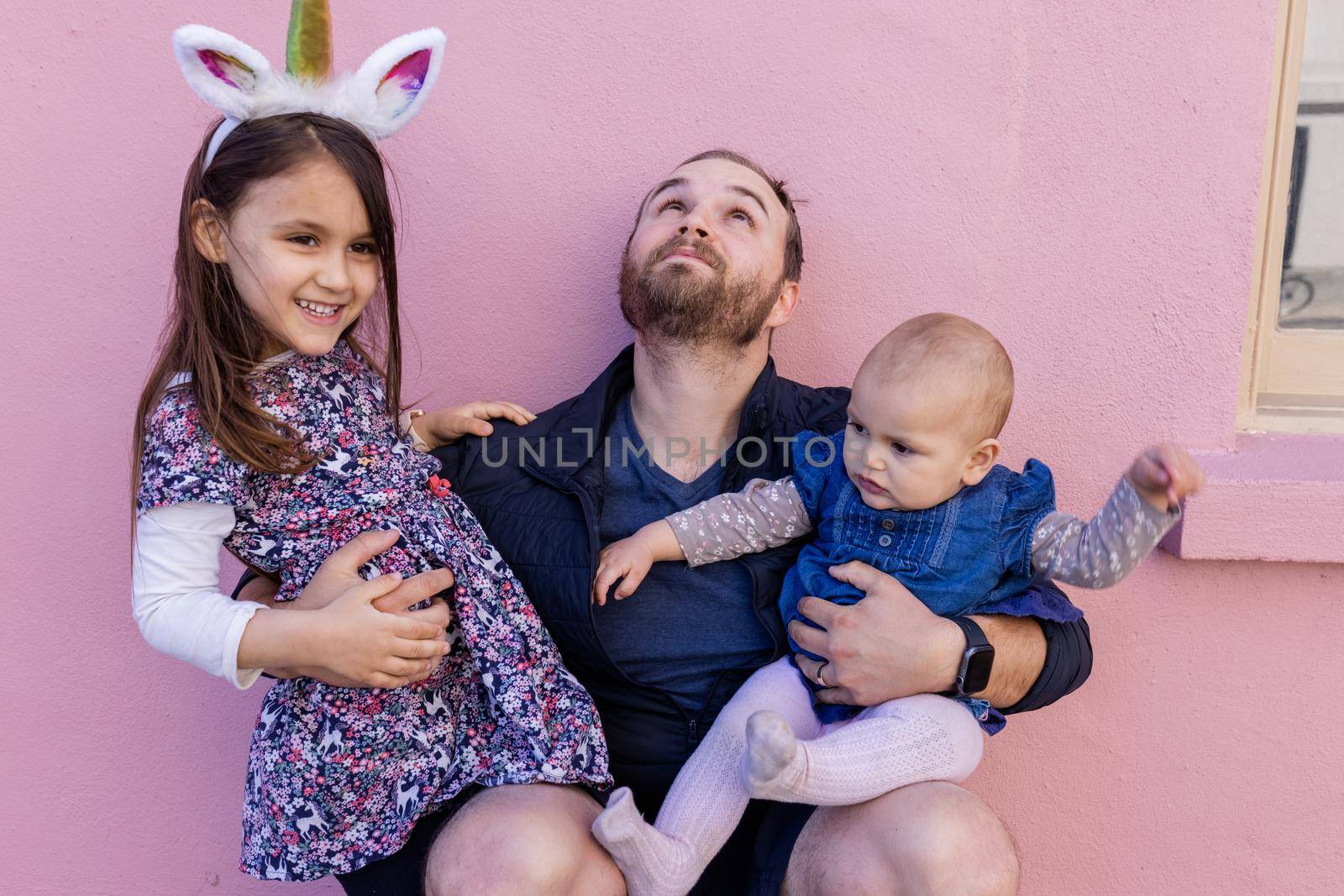 Portrait of young father looking up and hugging his adorable baby and older daughter in front of a pink wall. Bearded man in squatting position holding baby and little girl. Happy family outdoors