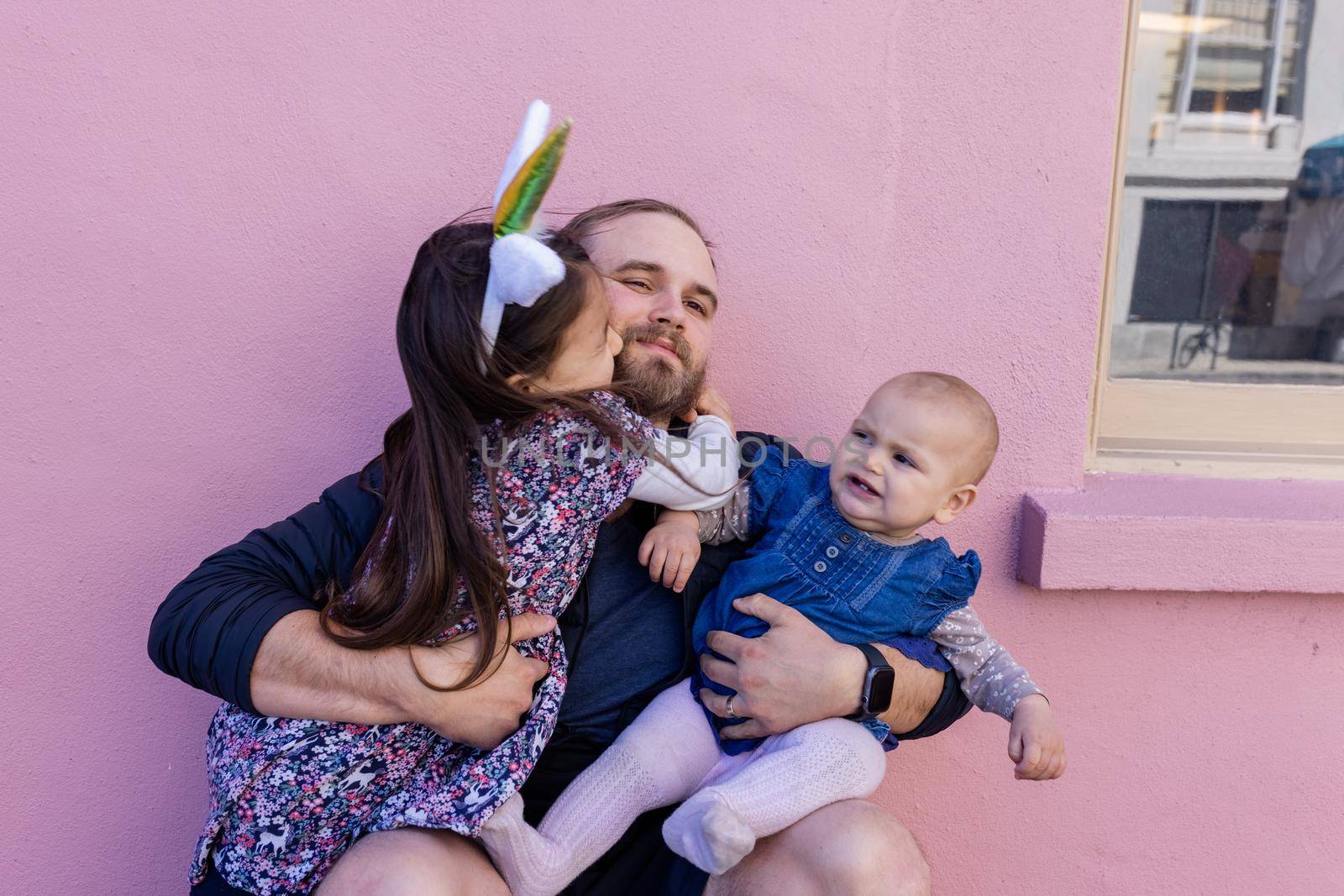 Portrait of little girl lovely hugging her father while holding cute baby in front of a pink wall. Adorable view of smiling bearded man holding joyful daughters. Happy family outdoors