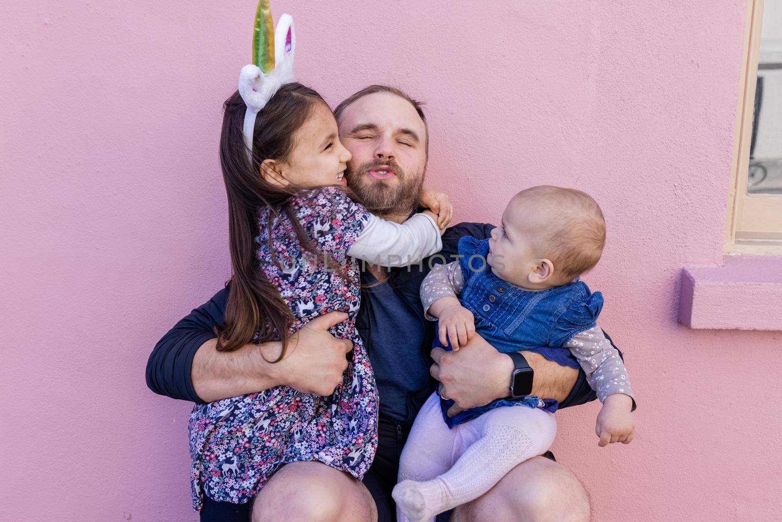Portrait of little girl lovely hugging her father while holding cute baby in front of a pink wall. Adorable view of smiling bearded man sitting and holding joyful daughters. Happy family outdoors