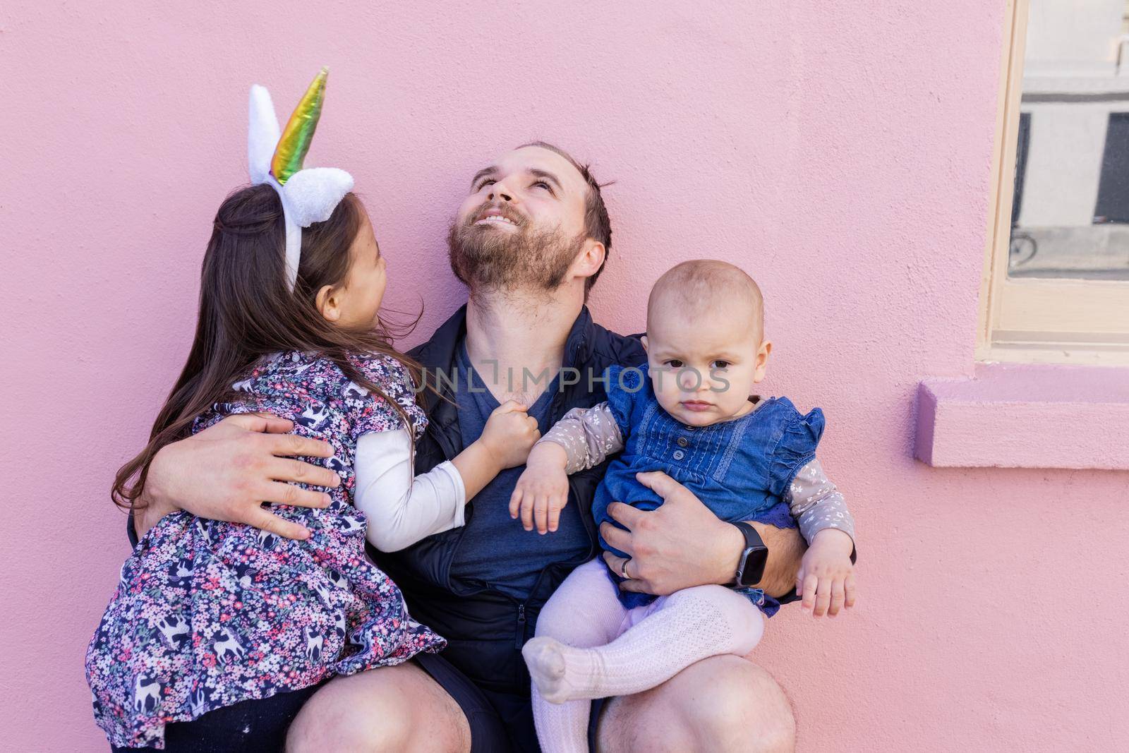 Portrait of young father looking up and hugging adorable baby and little girl in front of a pink wall. Bearded man sitting and holding joyful and cute daughters. Happy family outdoors