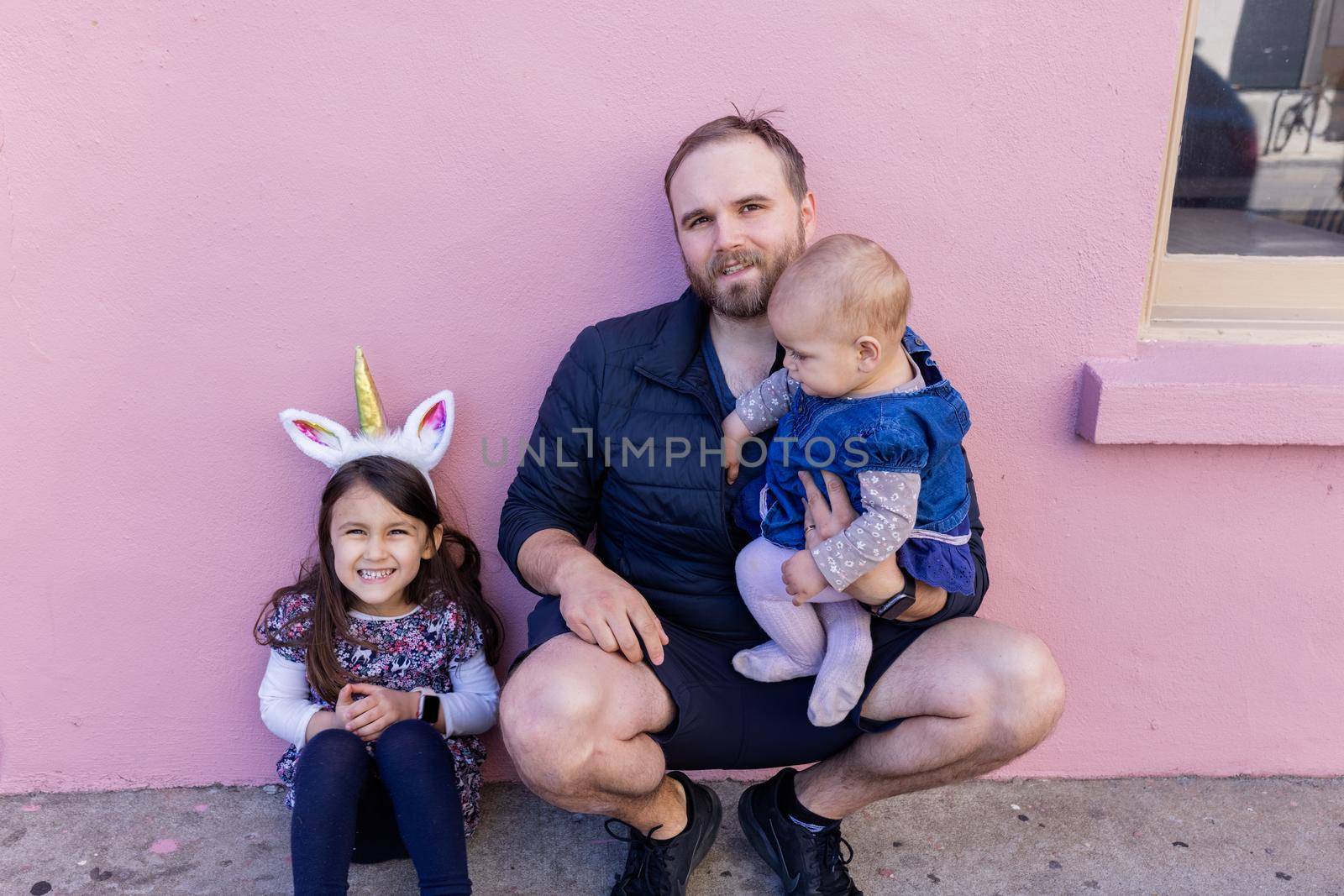 Portrait of young father with his adorable baby and older daughter in front of a pink wall. Cute child wearing unicorn headband next to baby and bearded man with pink background. Happy family outdoors