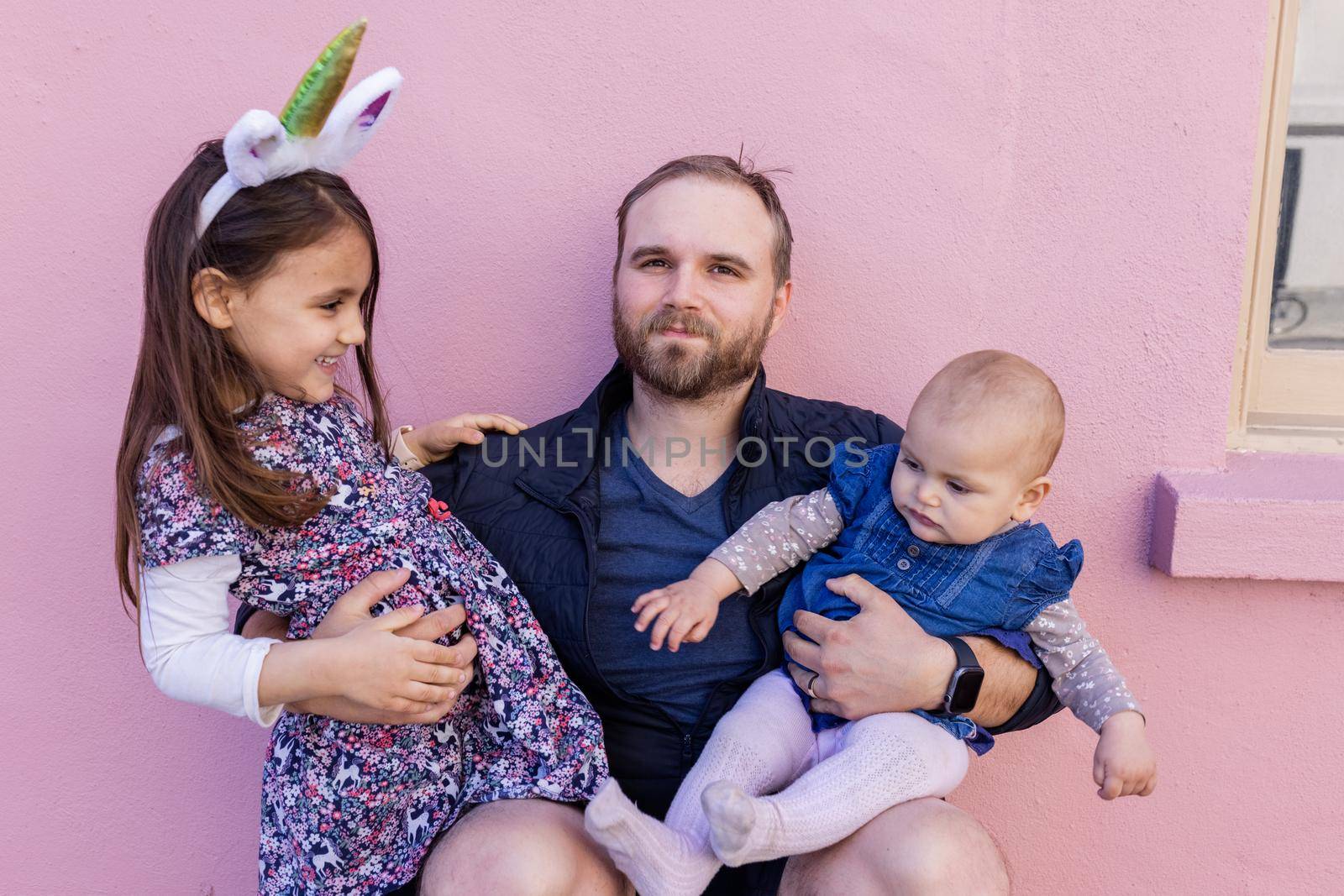 Portrait of young father hugging his adorable baby and older daughter in front of a pink wall. Bearded man sitting and holding joyful cute little girl and baby. Happy family outdoors