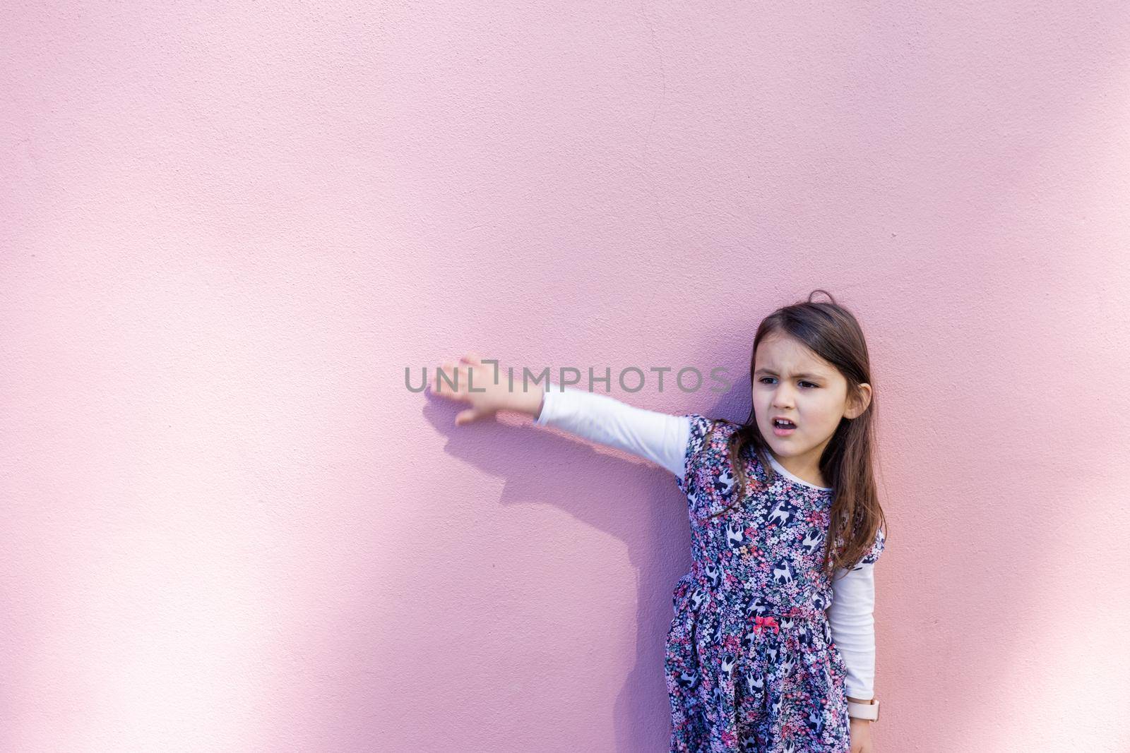 Adorable view of distressed-looking little girl wearing unicorn colorful dress with pink background. Portrait of cute young child touching a pink wall. Lovely kids outdoors