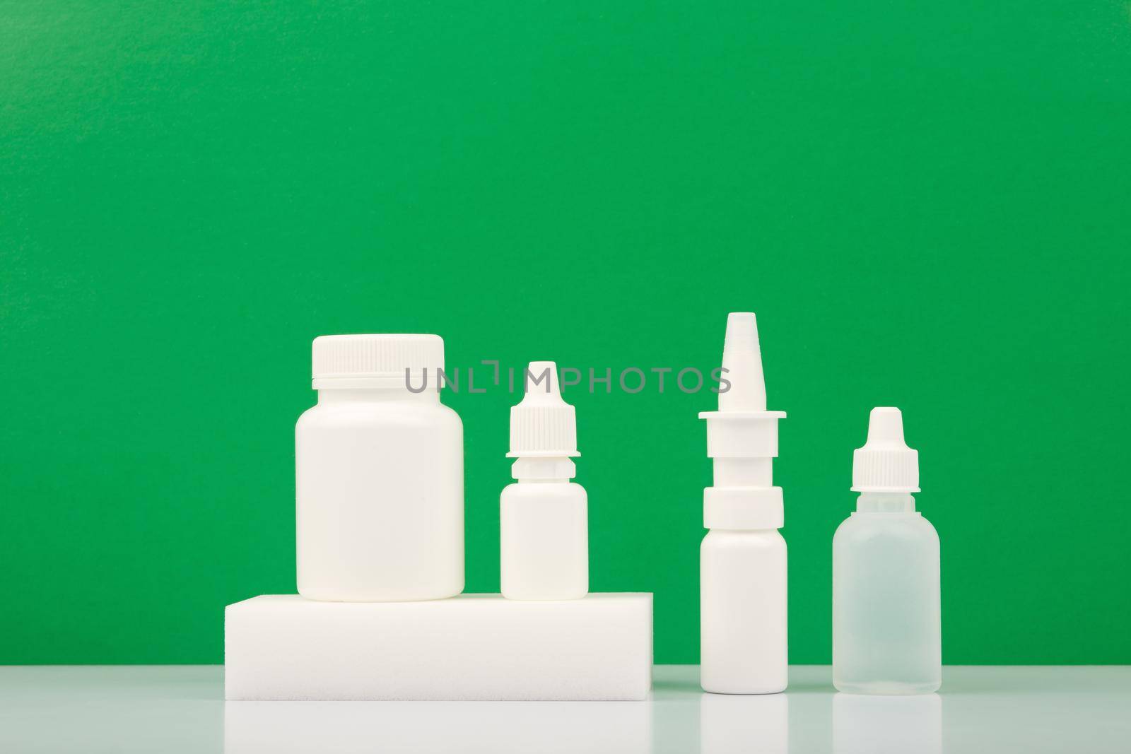 Medication pills and ear drops on podium and eye drops and nose spray against green background with copy space. Concept of health care and medications for treating cold-related diseases and flue