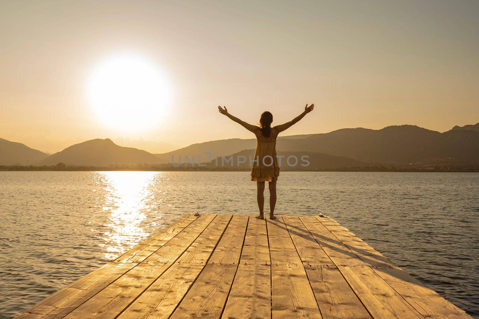 View from back of young woman on wooden pier in short dress and barefoot with open arms in contemplation of the sun setting among the mountains on lake or ocean sea. Orange vintage effect photography by robbyfontanesi