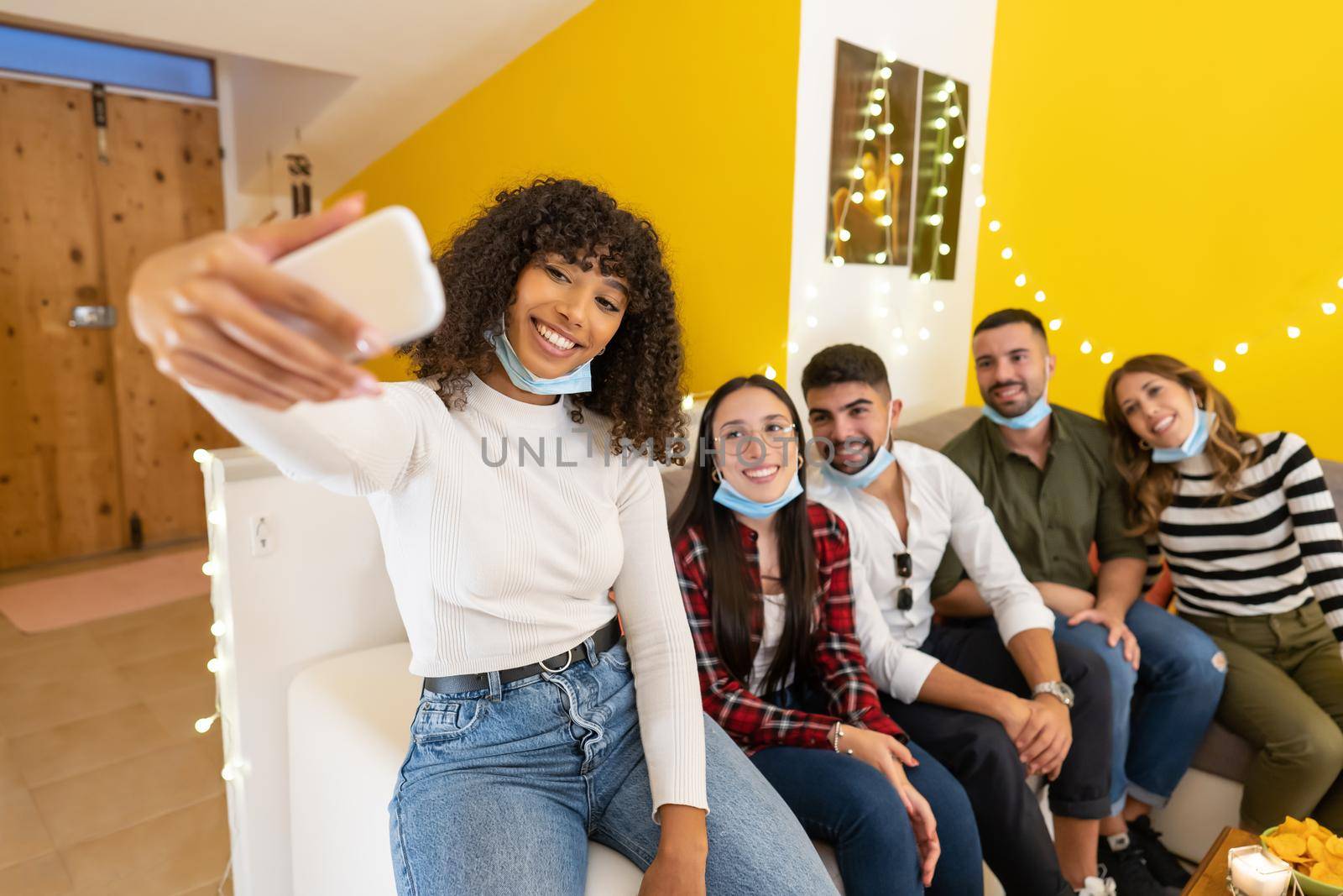 Group of young people at home wearing lowered protective medical mask posing for selfie sitting on a couch at home party, Afro american girl using smartphone to share on social network happy moments