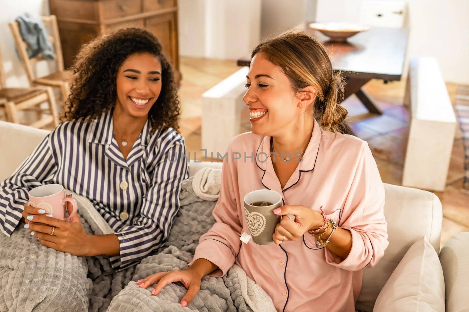 Elegance, beauty and happiness: multiracial young women couple having fun wearing pajama sitting on the sofa drink cup of tea. Afro american curly woman laughing at home with her best caucasian friend