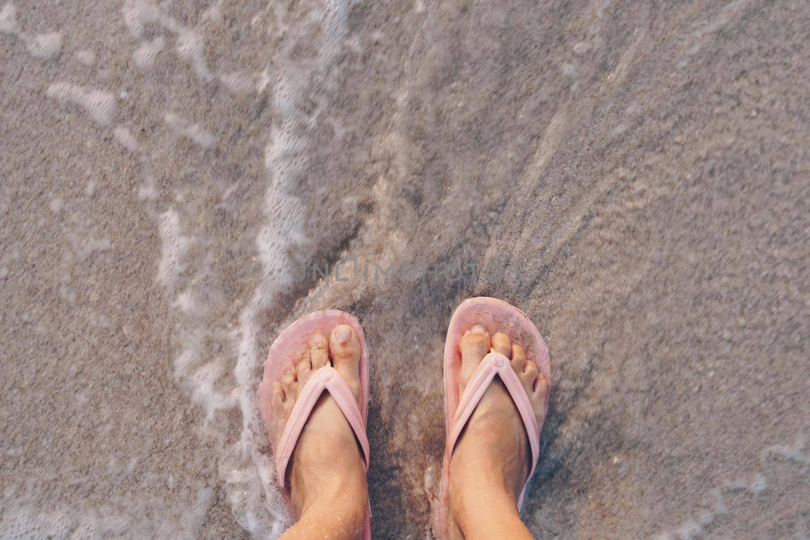 Woman feet wear slippers stand on sand tropical beach with sea water splash. by Suwant