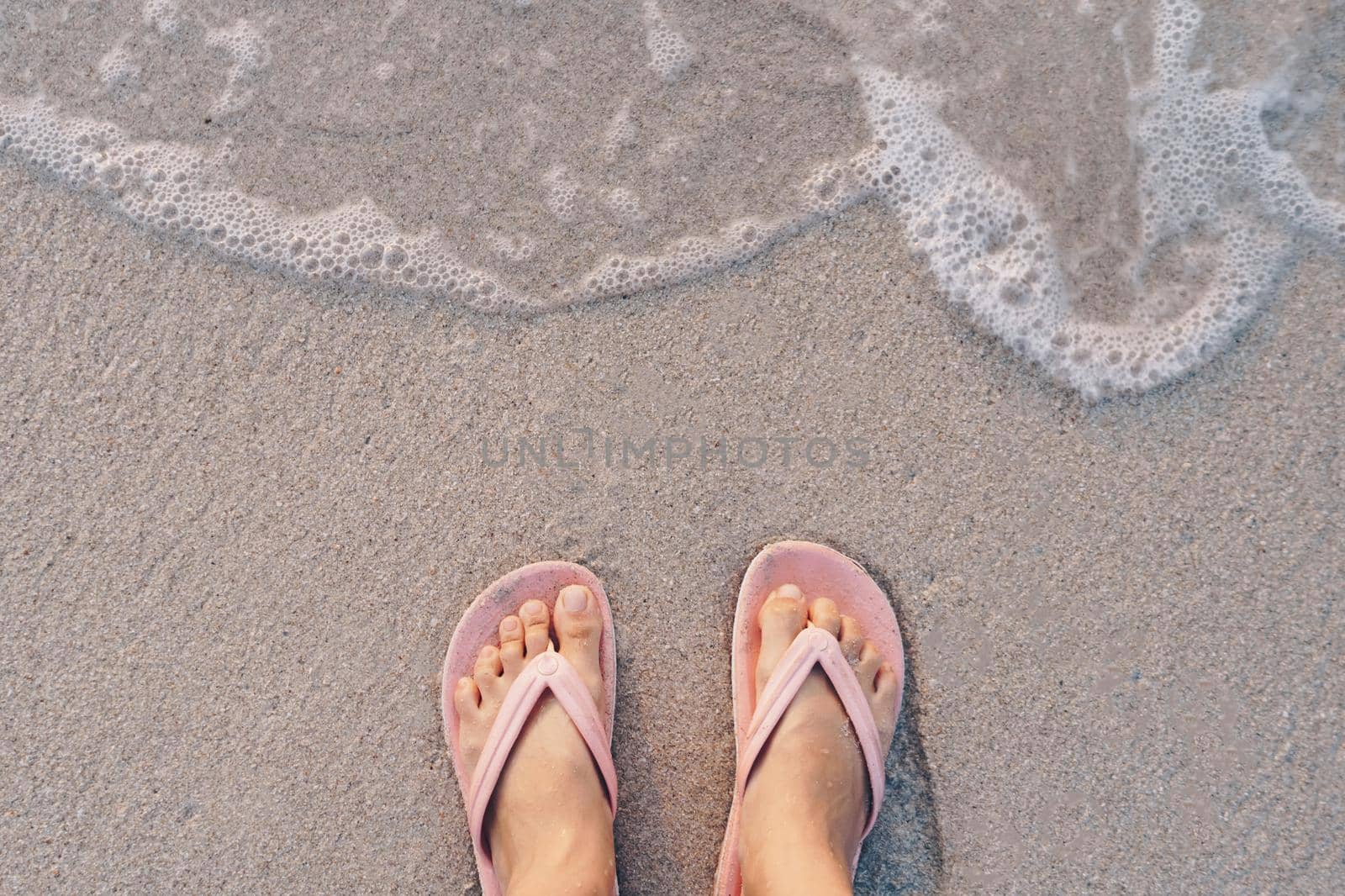 Woman feet wear slippers stand on sand tropical beach with sea water splash background.