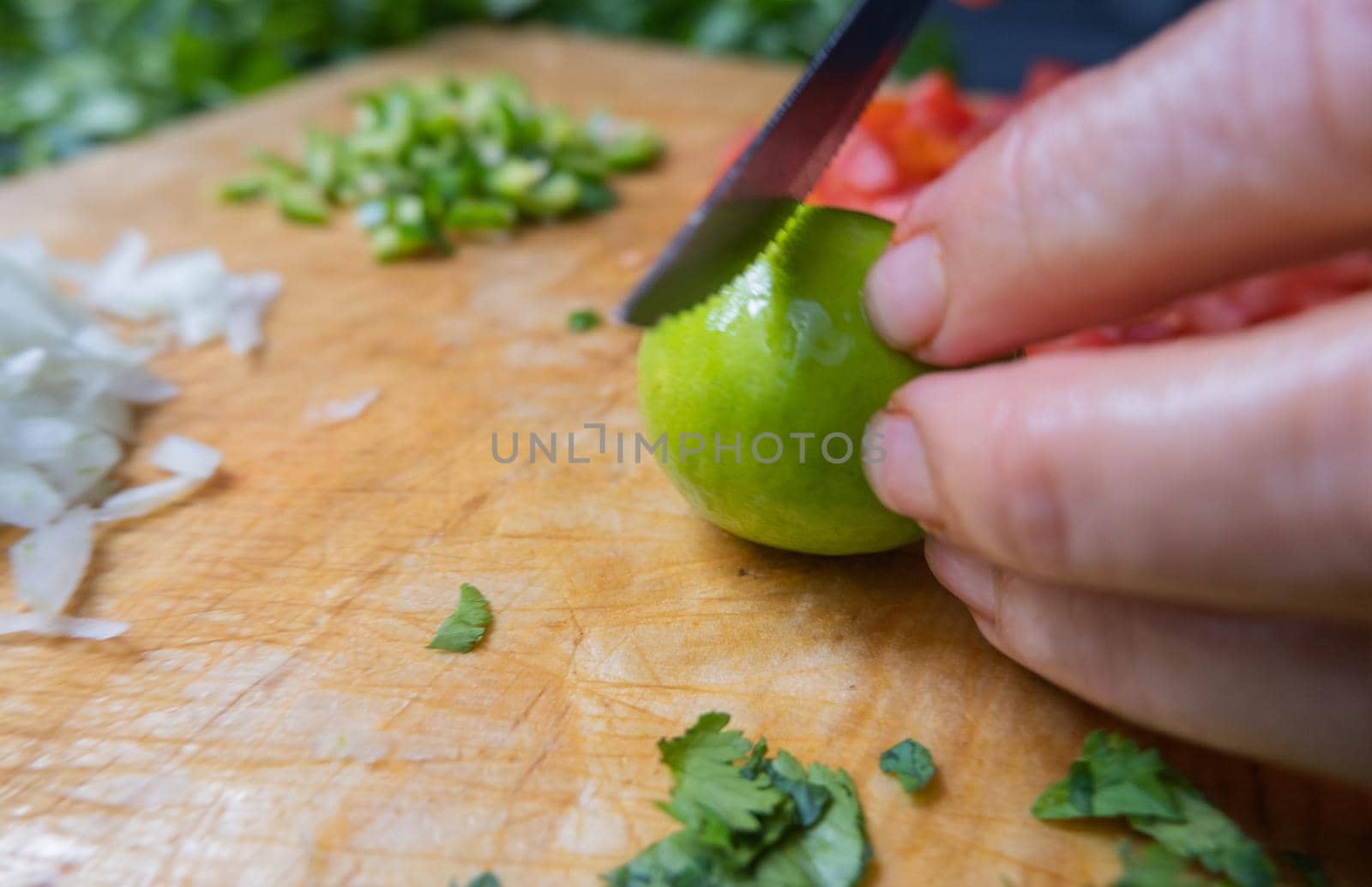 Close-up of hands slicing lime on wooden cutting board with chopped tomatoes, onions, and peppers. Person cutting fresh vegetables into pieces on wood surface. Traditional sauce preparation