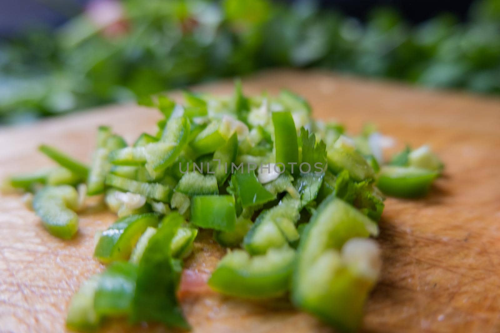 Close-up of chopped chili pepper on wooden cutting board with blurry coriander as background. Fresh green vegetables and herbs cut into pieces above wood surface. Traditional sauce preparation