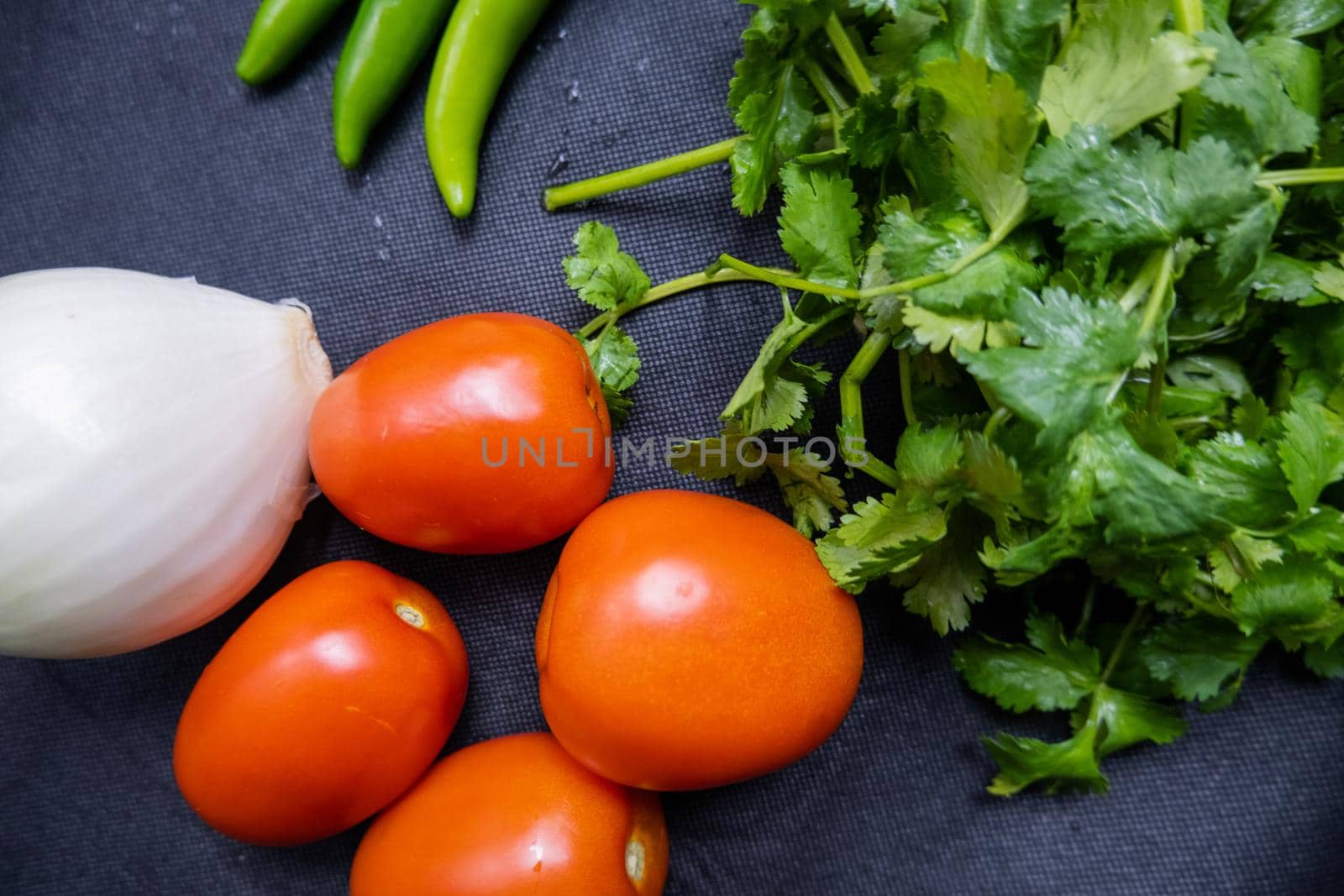 Top view of four tomatoes, green chili peppers, onion, and coriander on table. Fresh and aromatic vegetables and herbs on dark blue surface. Healthy meal preparation