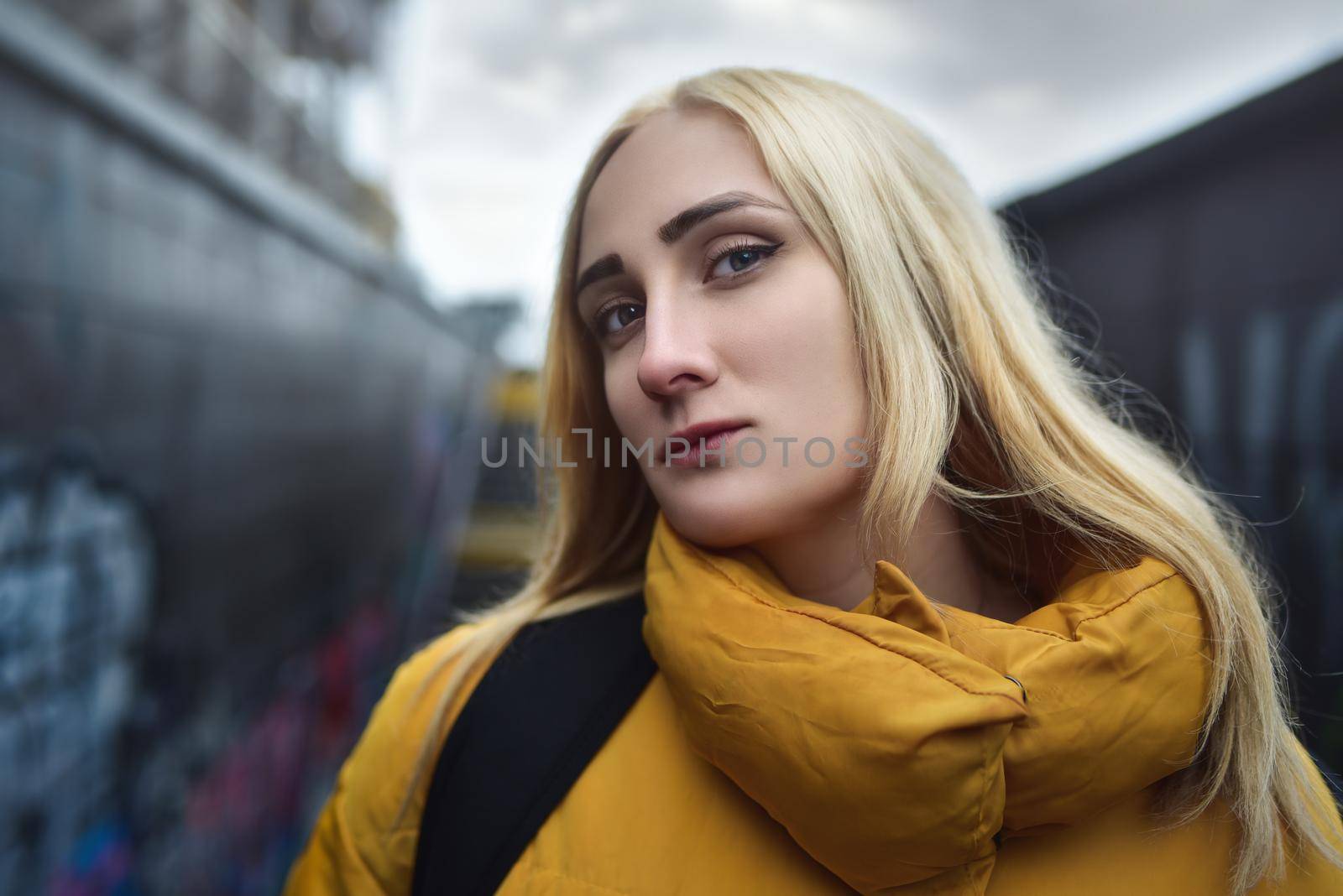 Close up portrait of blonde hipster teen girl making selfie, She is wearing yellow jacket and backpack, walking outdoors throught underground passage and making selfie