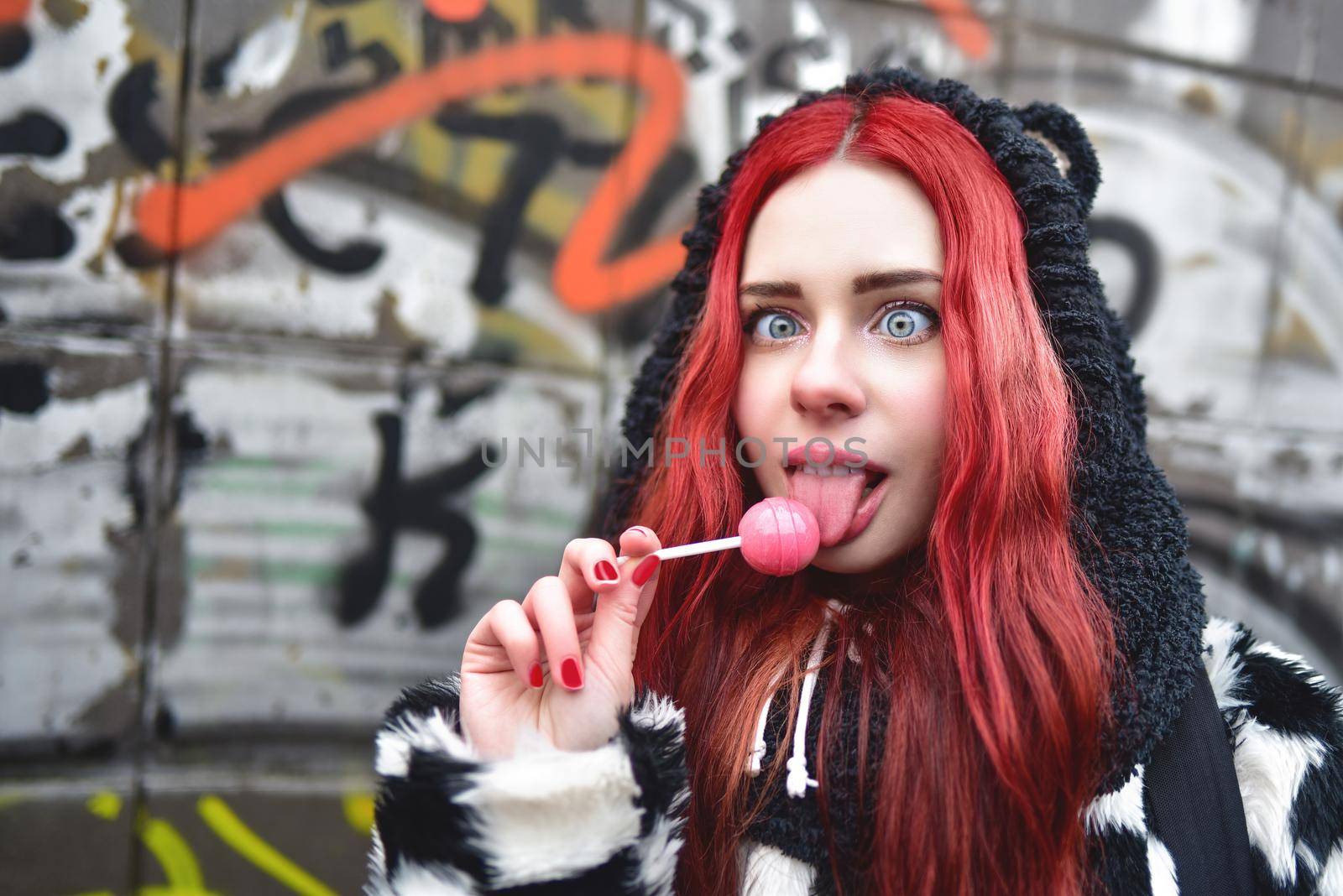 Portrait of a young beauty teen girl in a hoodie with a delicious pink lollipop, posing against a wall of graffiti. Positive Emotions. Urrban style