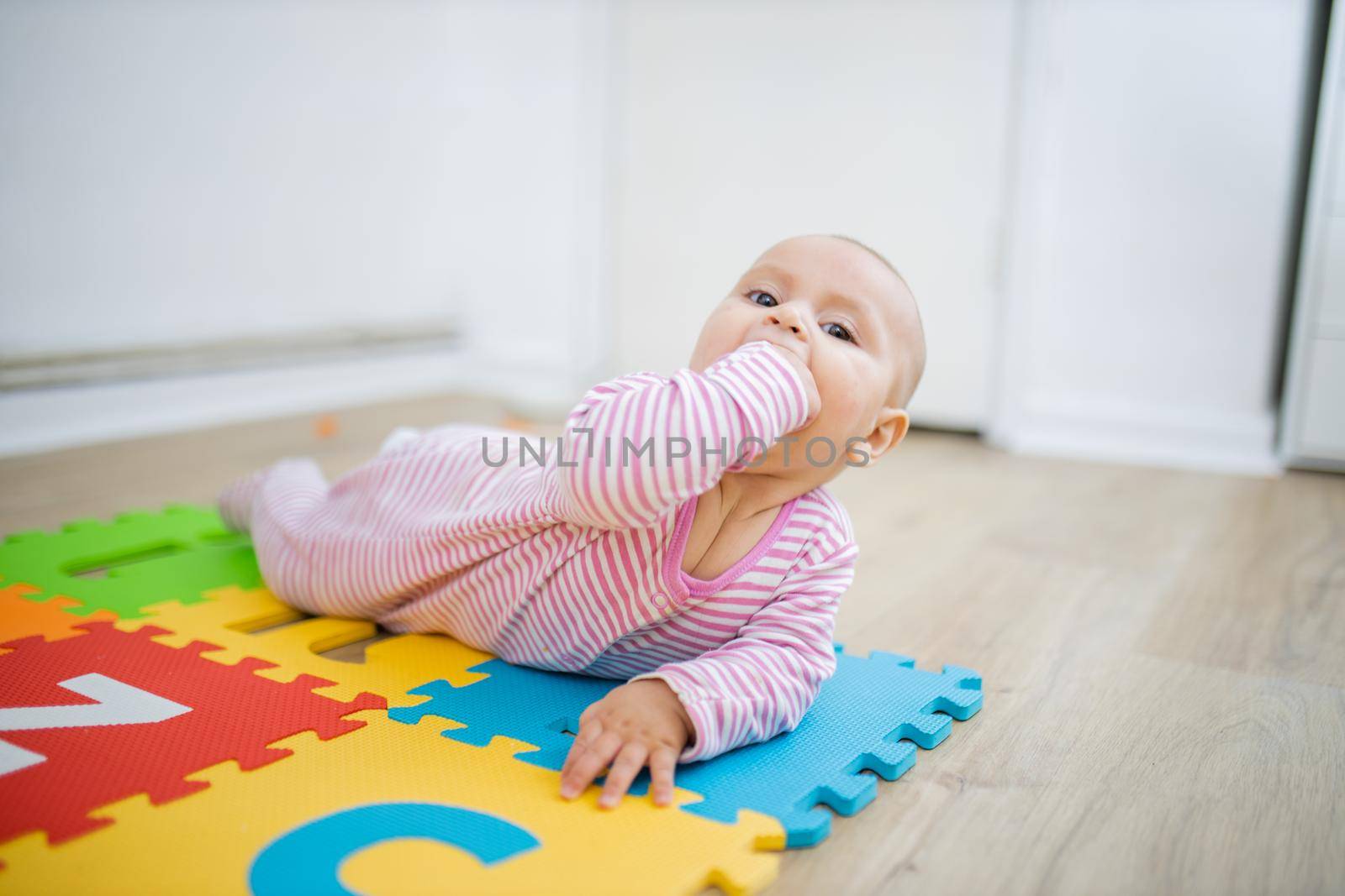 Adorable baby lying face down on colorful children mat with her fist in her mouth. Portrait of curious baby playing on the floor. Happy babies having fun