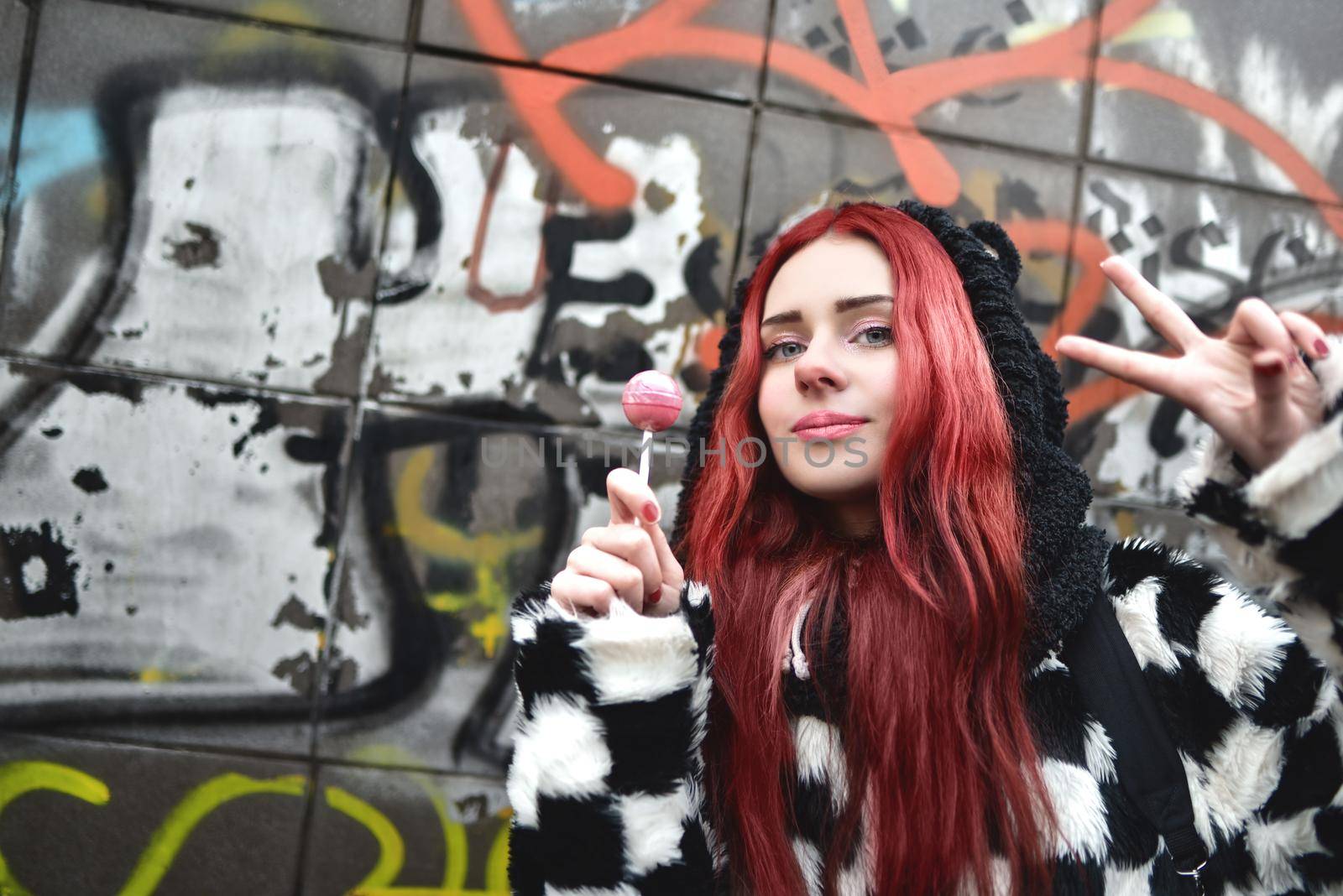 Portrait of a young beauty in a hoodie with a delicious pink lollipop, posing against a wall of graffiti. Positive Emotions. by Nickstock