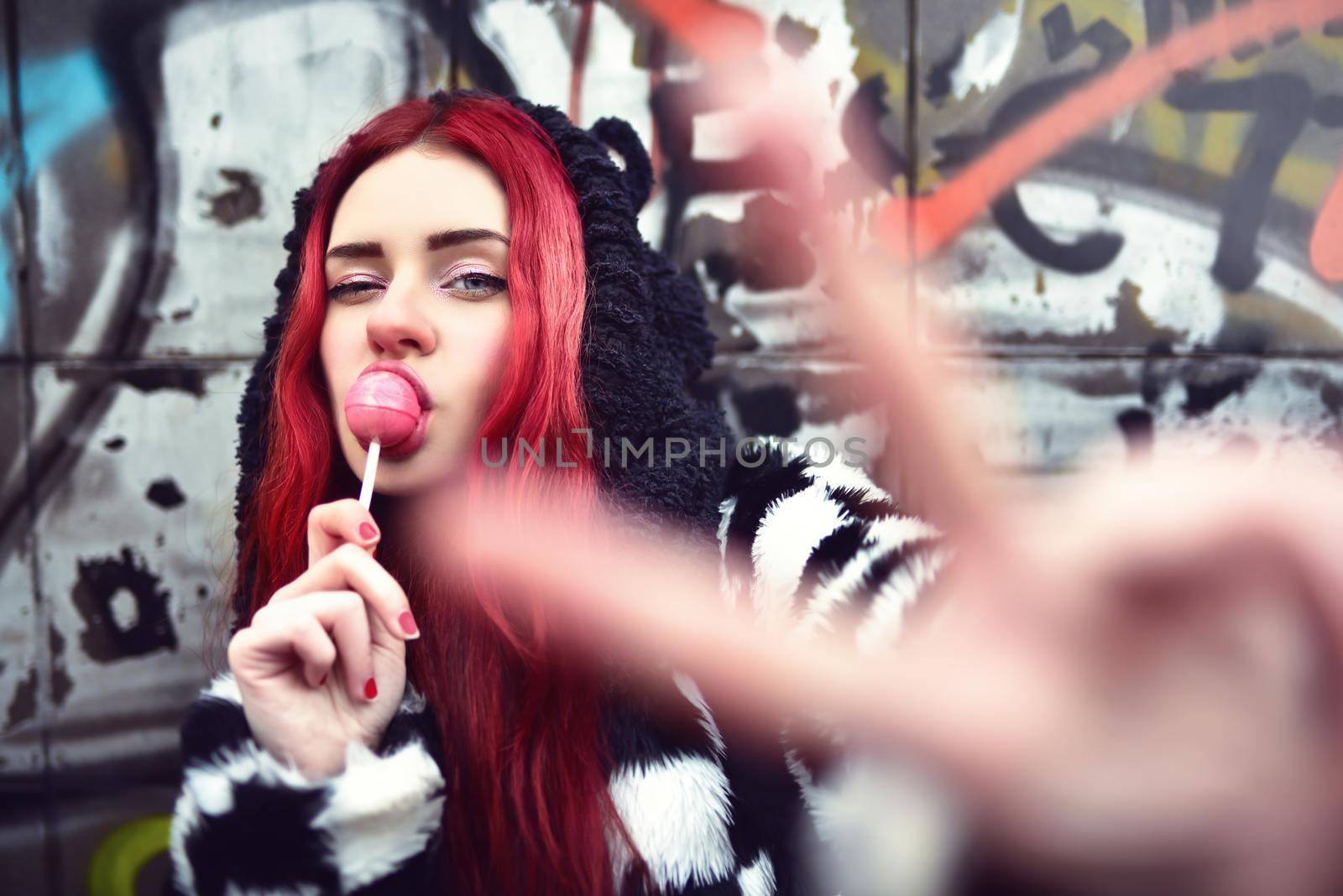 Portrait of a young beauty in a hoodie with a delicious pink lollipop, posing against a wall of graffiti. Positive Emotions. by Nickstock