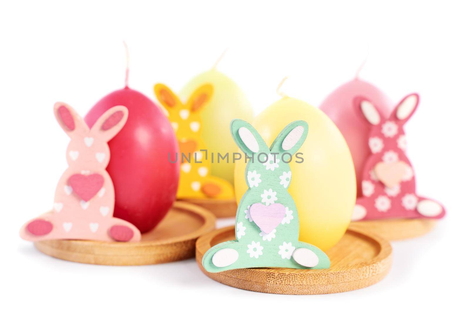 Close up of small Easter Bunnies next to egg shaped candles in pastel colors, isolated on white background. Fun and colorful Easter celebration concept.