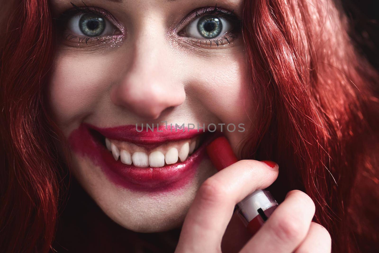 Portrait of crazy-looking teen girl with red hair she is smearing red lipstick on her face, horror concept. halloween time. Fear and nightmare. close view