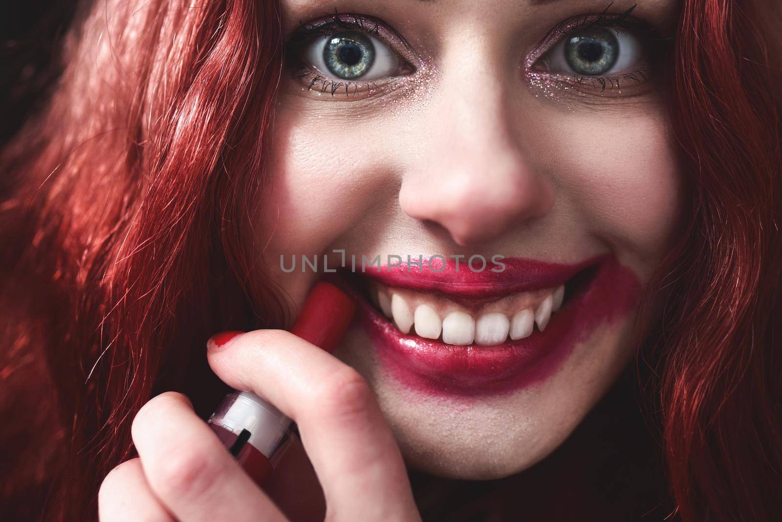 Portrait of crazy-looking teen girl with red hair she is smearing red lipstick on her face, horror concept. halloween time by Nickstock