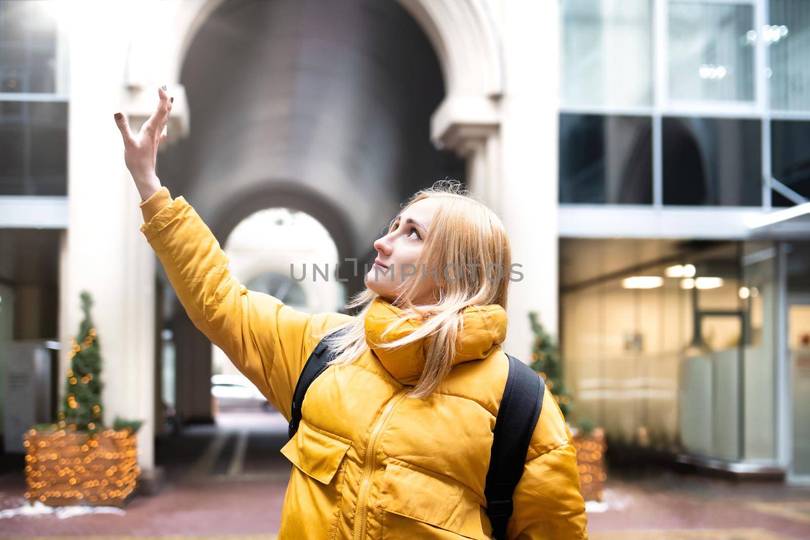 Cheerful attractive young blonde woman wearing yellow jacket with backpack walking in the city by Nickstock