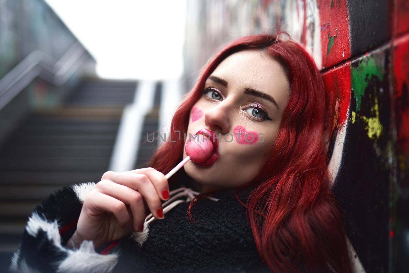 A glamorous teenage girl with red hair licks a sweet strawberry candy near a wall of graffiti. by Nickstock