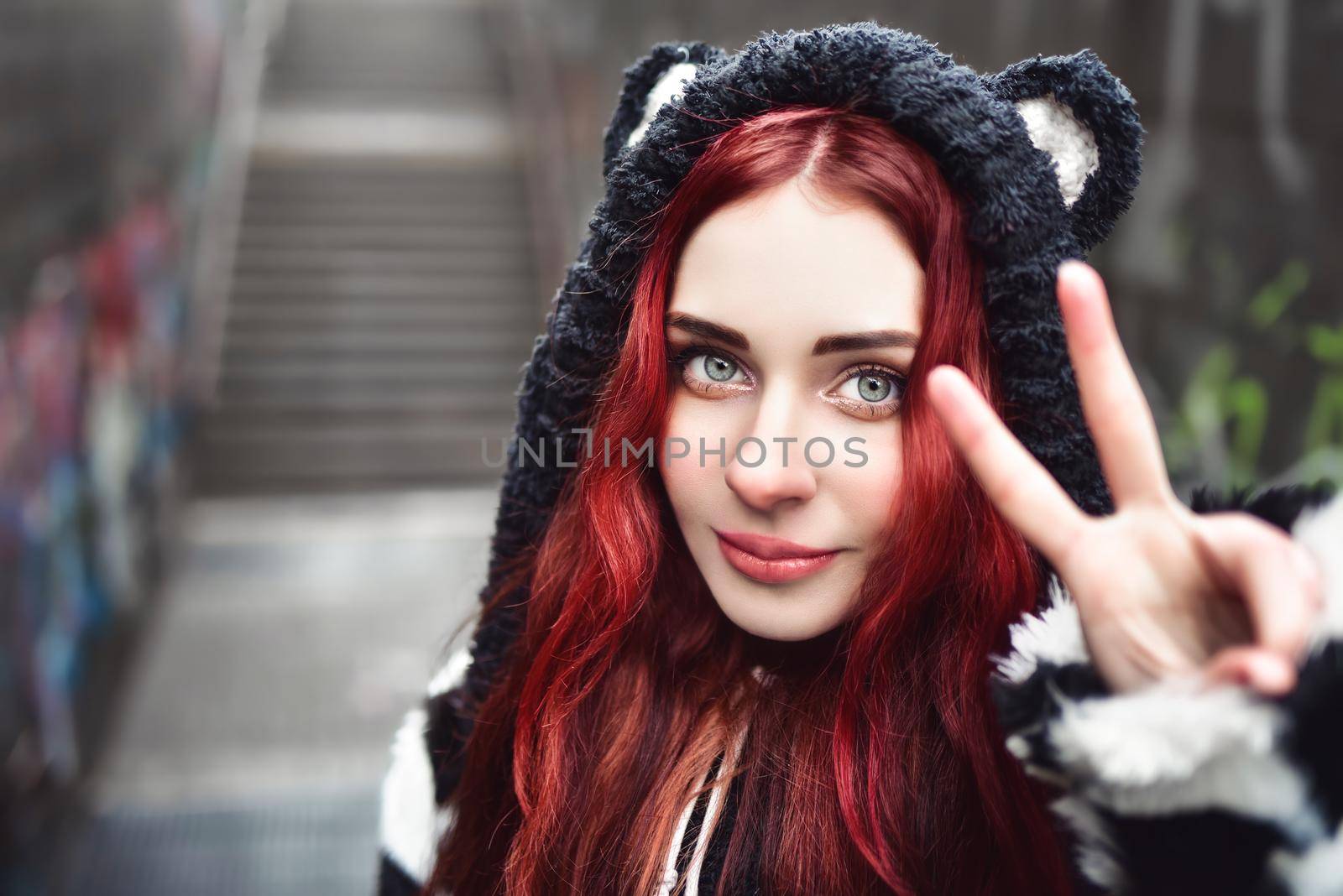 Close up portrait of cheerful playful girl, who is gesturing peace sign and smiles. She is wearing hoody, walking outdoors throught underground passage