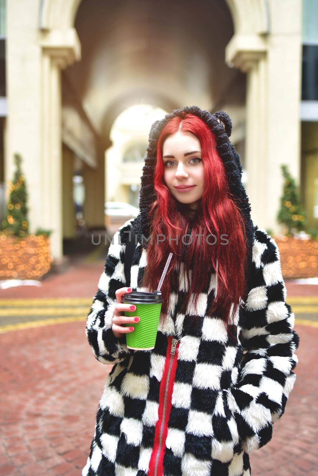 Near portrait of a teen girl with red hair in warm clothes standing outside on a cold day with a cup of coffee in her hands and looks into the camera. Cute girl walking with coffee in her hands.