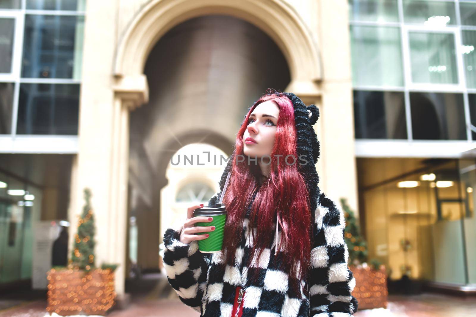a portrait of a teen girl with red hair in warm clothes walking outside on a cold day with a cup of coffee in her hands and looking up. Cute girl walking with coffee in her hands. by Nickstock