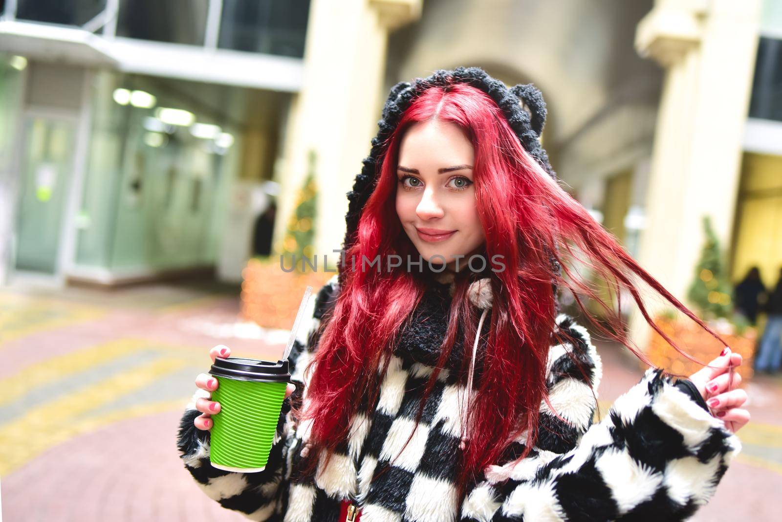 Near portrait of a teen girl with red hair in warm clothes standing outside on a cold day with a cup of coffee in her hands and looks into the camera. Cute girl walking with coffee in her hands and playing with own hair.