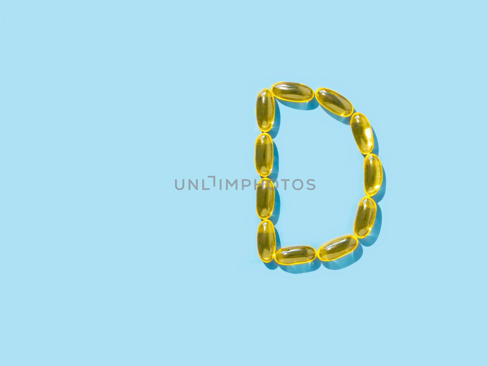 Vitamin D3, Omega or Evening Primrose Oil gel capsules on blue background. Top view or flat lay. Letter D made from yellow liquid capsules with D3 nutritional supplement oil. Copy space left.