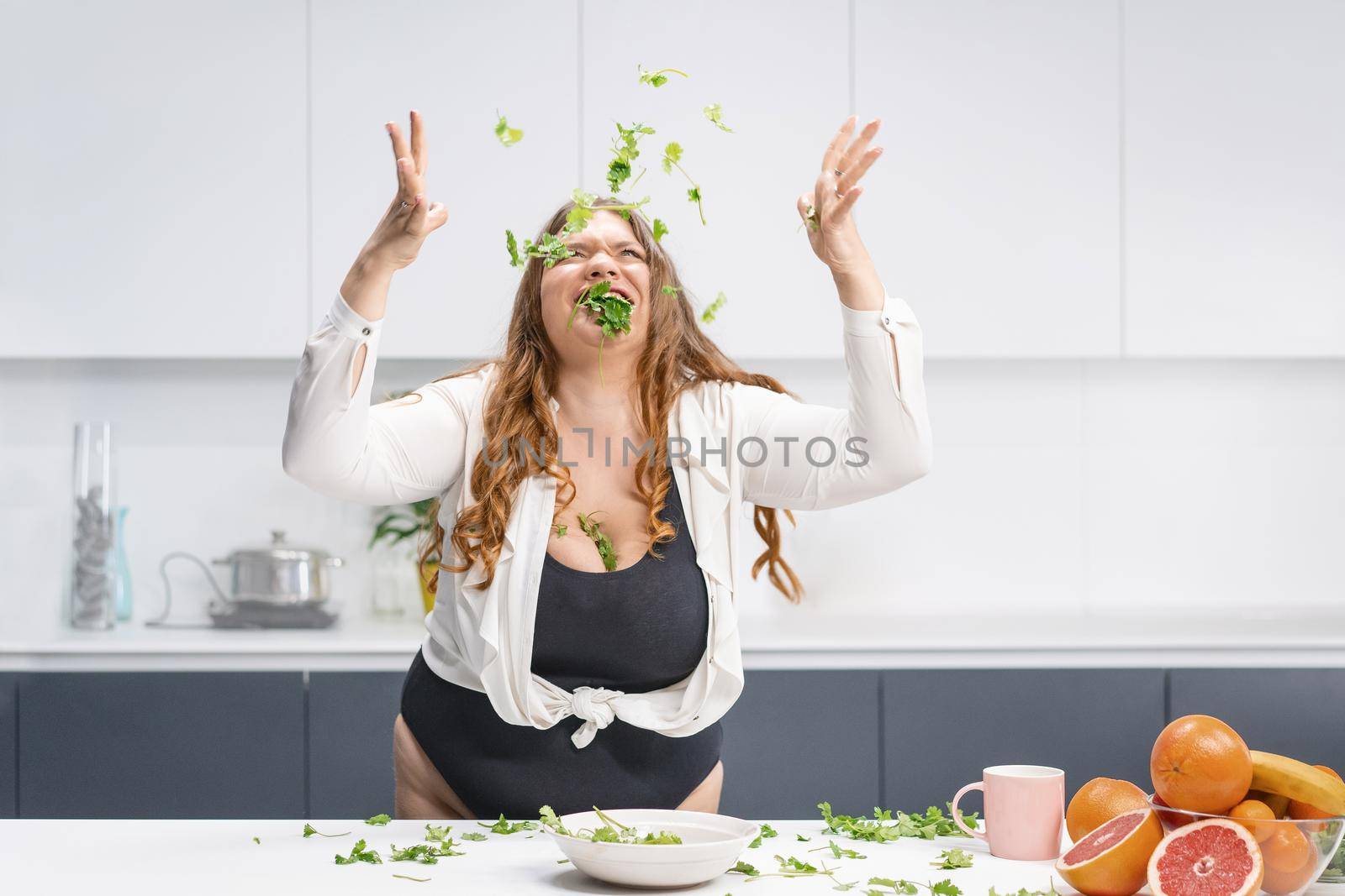 Happy chubby girl scattering spinach leaves all over the table. Overweight girl happy loosing weight eating fresh salad. Curvy body young woman with long blond hair. Dieting and nutrition concept by LipikStockMedia