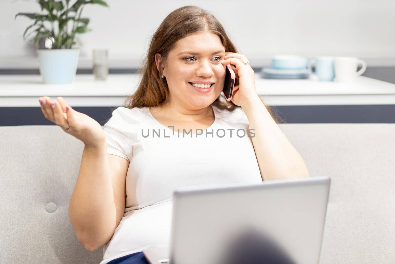 Talking on the phone with nutritionist looking at the program on a laptop young fat girl staying at home during quarantine. Self isolation as prevention. Work distantly concept by LipikStockMedia