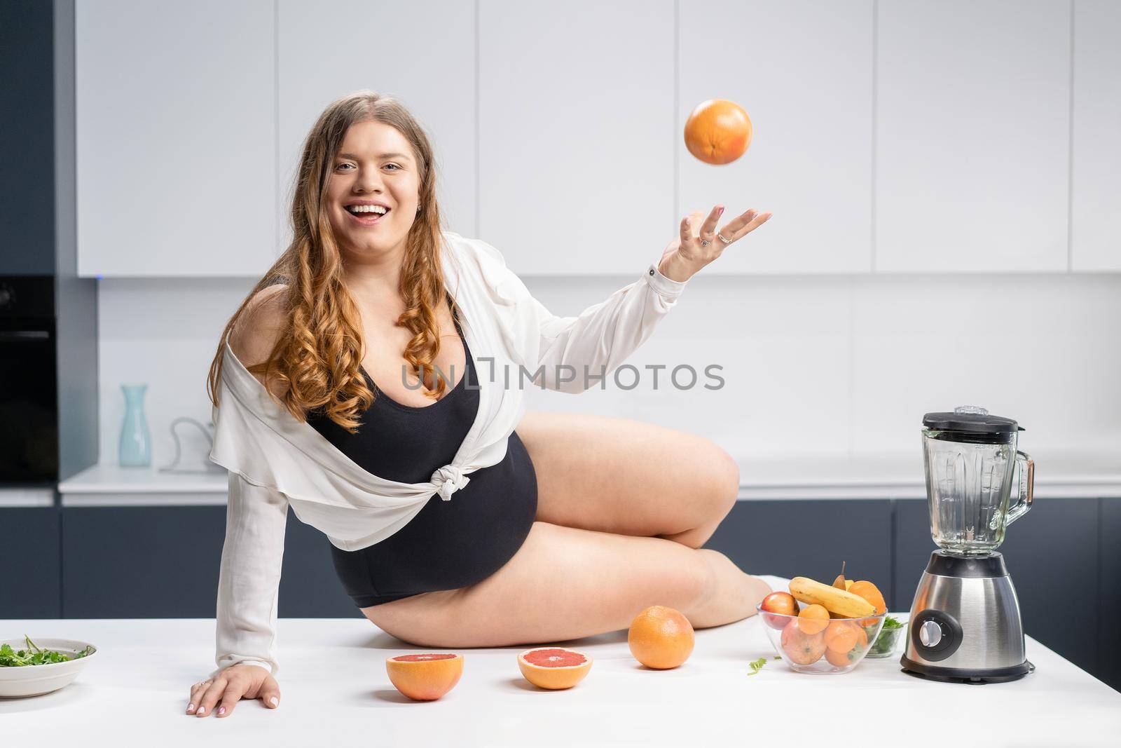Girl with obese problem. Young sexy chubby white girl in black swimsuit, white shirt at modern kitchen table. juggling fruits in hand. Trying to loose weight fast. Fat barefoot girl low carb diet.