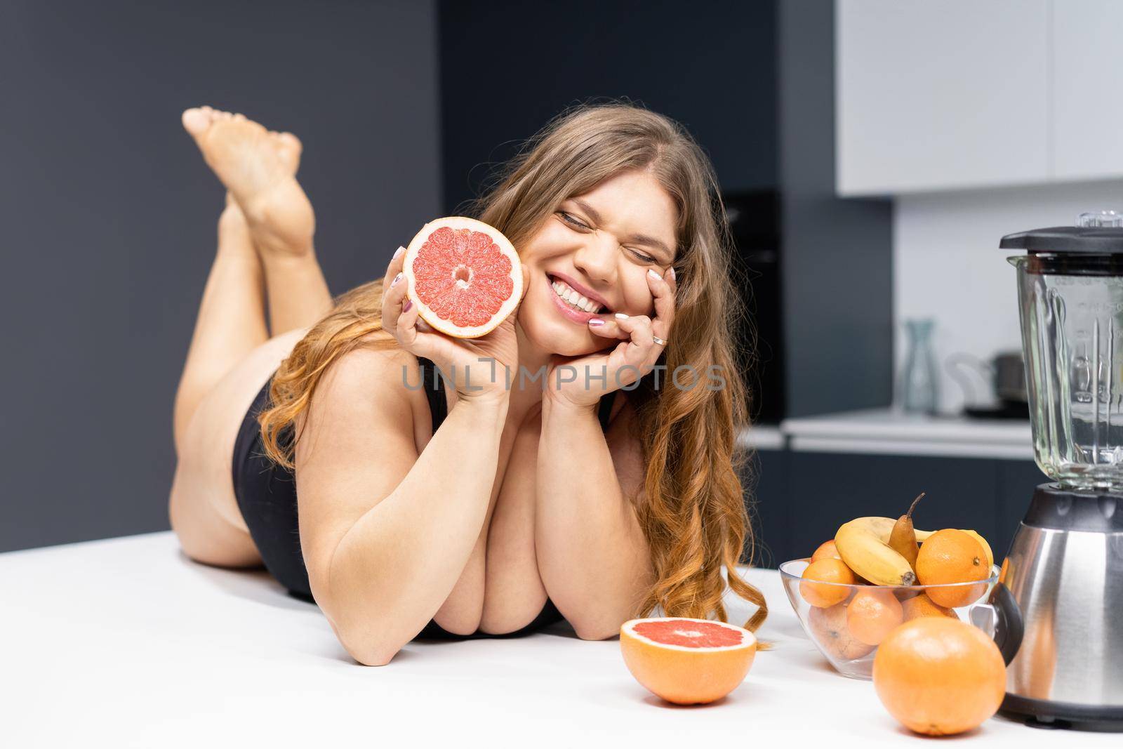 Young sexy chubby white girl in black swimsuit, white shirt at modern kitchen table. juggling fruits in hand. Trying to loose weight fast. Girl with obese problem. Fat barefoot girl low carb diet by LipikStockMedia