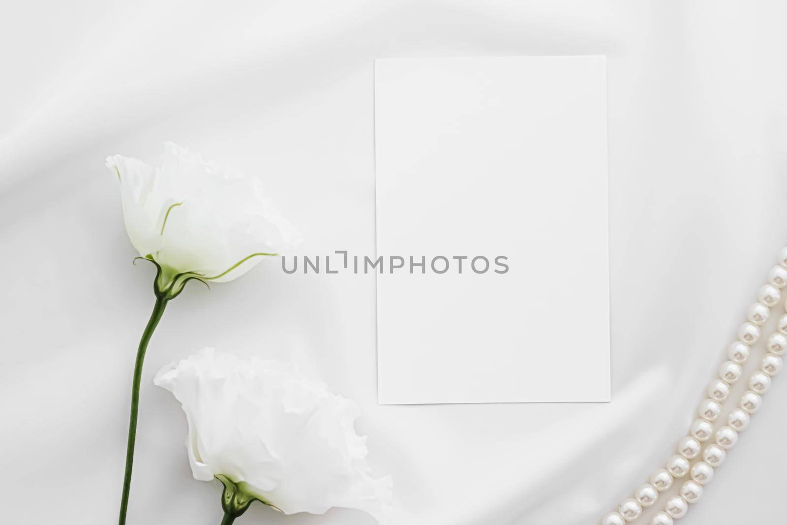 Wedding invitation, white rose flowers and pearls on silk fabric as bridal flatlay background, blank paper greeting card and holiday branding, flat lay design concept