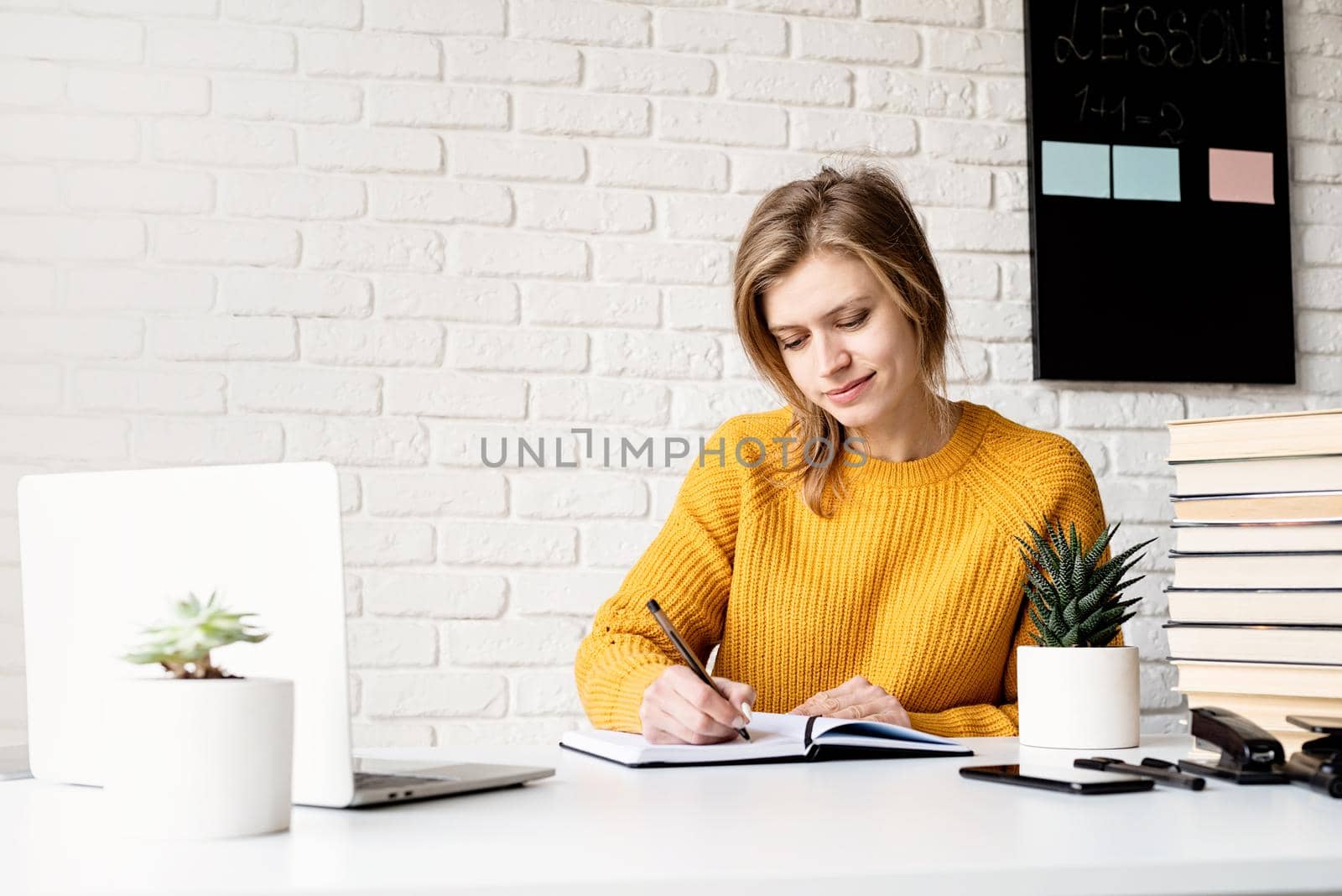 Distance learning. E-learning. Young smiling woman in yellow sweater studying online using laptop writing in notebook
