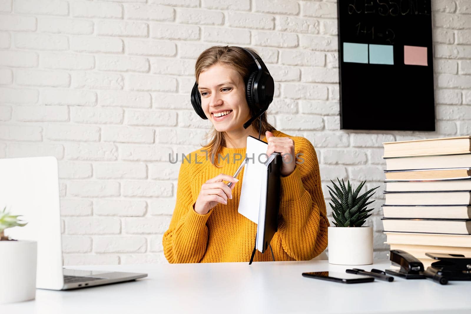 Distance learning. E-learning. Young smiling woman in yellow sweater and black headphones studying online using laptop writing in notebook