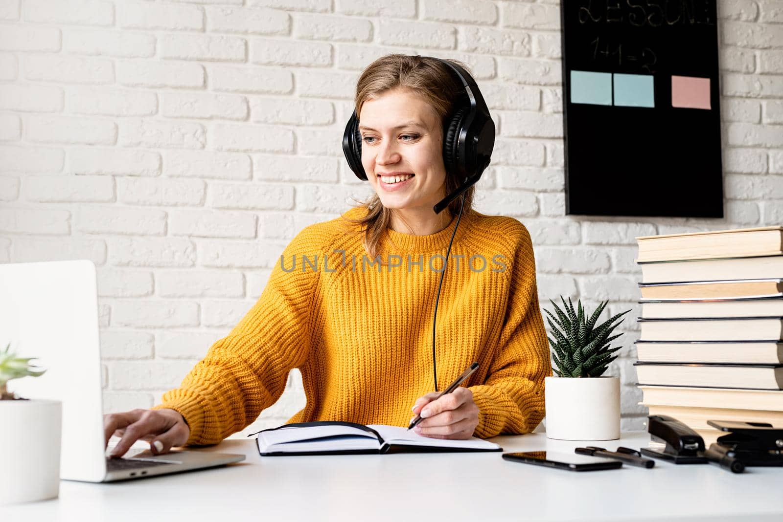 Young smiling woman in black headphones studying online using laptop writing in notebook by Desperada