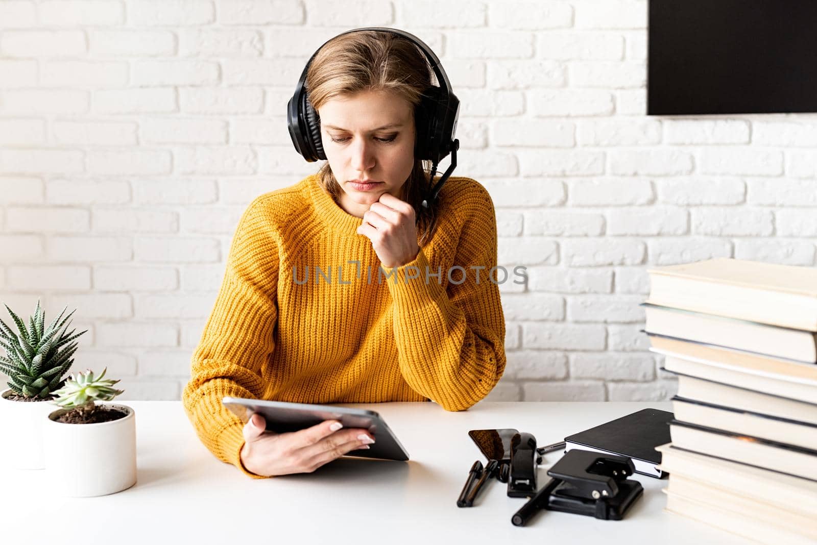 Distance learning. E-learning. Young thoughtful woman in yellow sweater and black headphones studying online using digital tablet